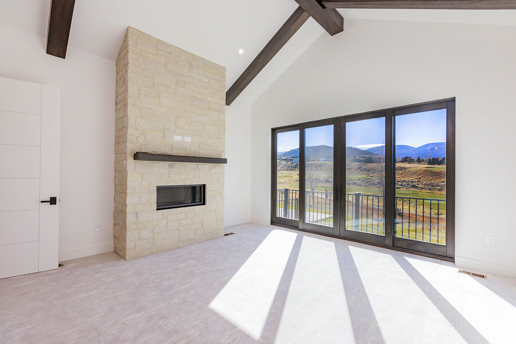 This is a bright and spacious living room featuring a stone fireplace, dark wood ceiling beams, and a large sliding glass door that opens to a balcony with views of a golf course and mountains. The room has white walls and light-colored carpet, creating a modern and airy feel. The perspective is from the corner of the room, showcasing the fireplace and the view.