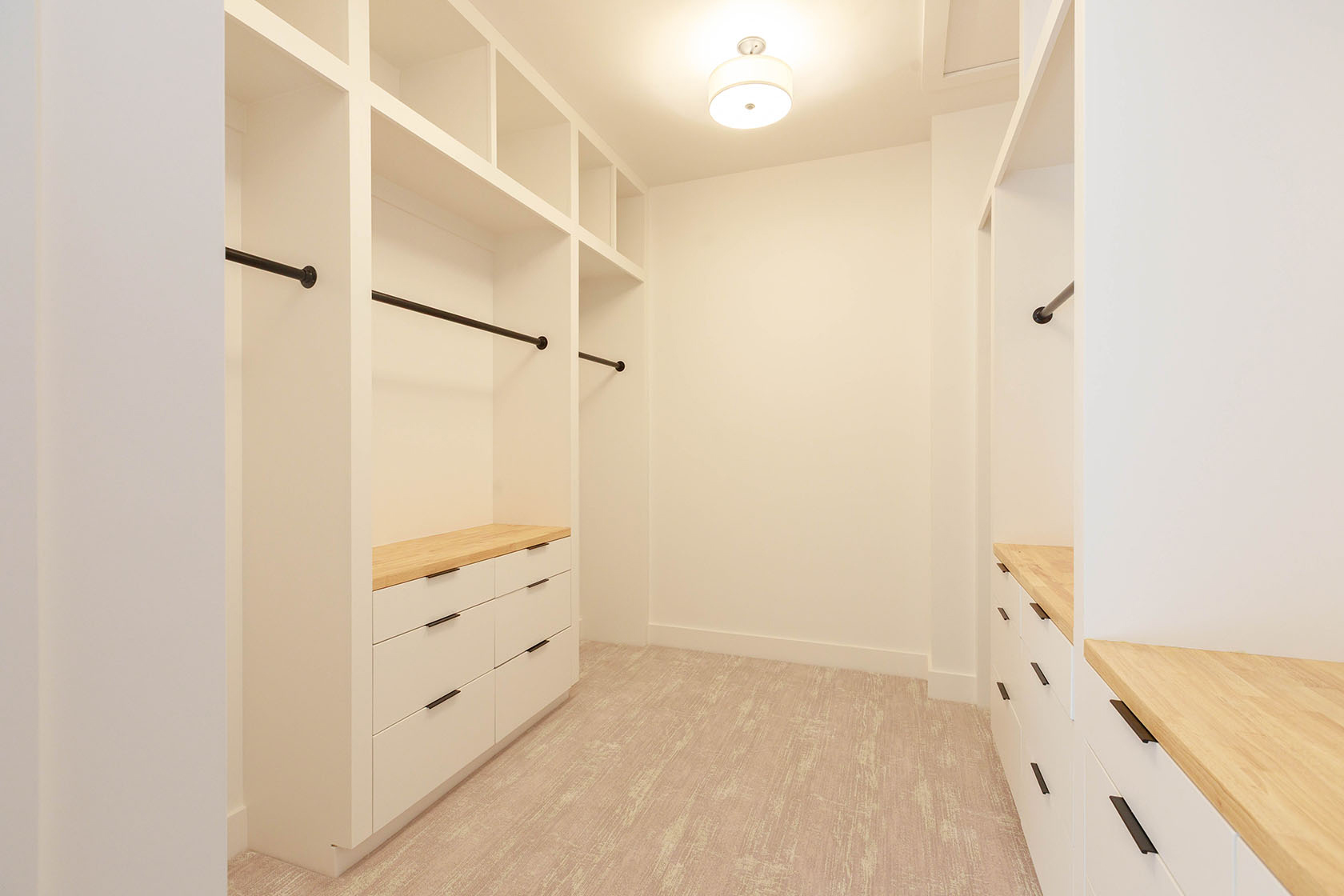 This is a well-organized walk-in closet featuring custom white shelving and drawers with wooden countertops. Black rods provide ample hanging space, and the neutral-toned carpet adds warmth to the space. The closet is brightly lit by a ceiling light, creating a clean and inviting atmosphere.