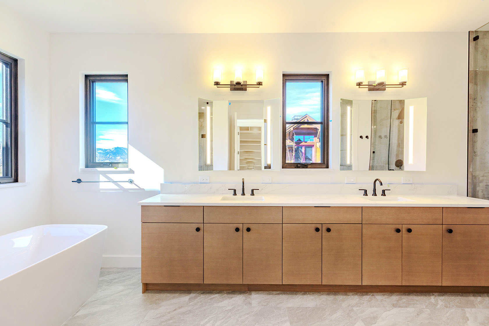 This is a bright and modern primary bathroom featuring a long, light wood vanity with double sinks and a white countertop. Large mirrors with integrated lighting are above the sinks, and two windows provide natural light and views. A freestanding white bathtub is visible on the left, and the flooring is a light gray tile, creating a clean and luxurious space.