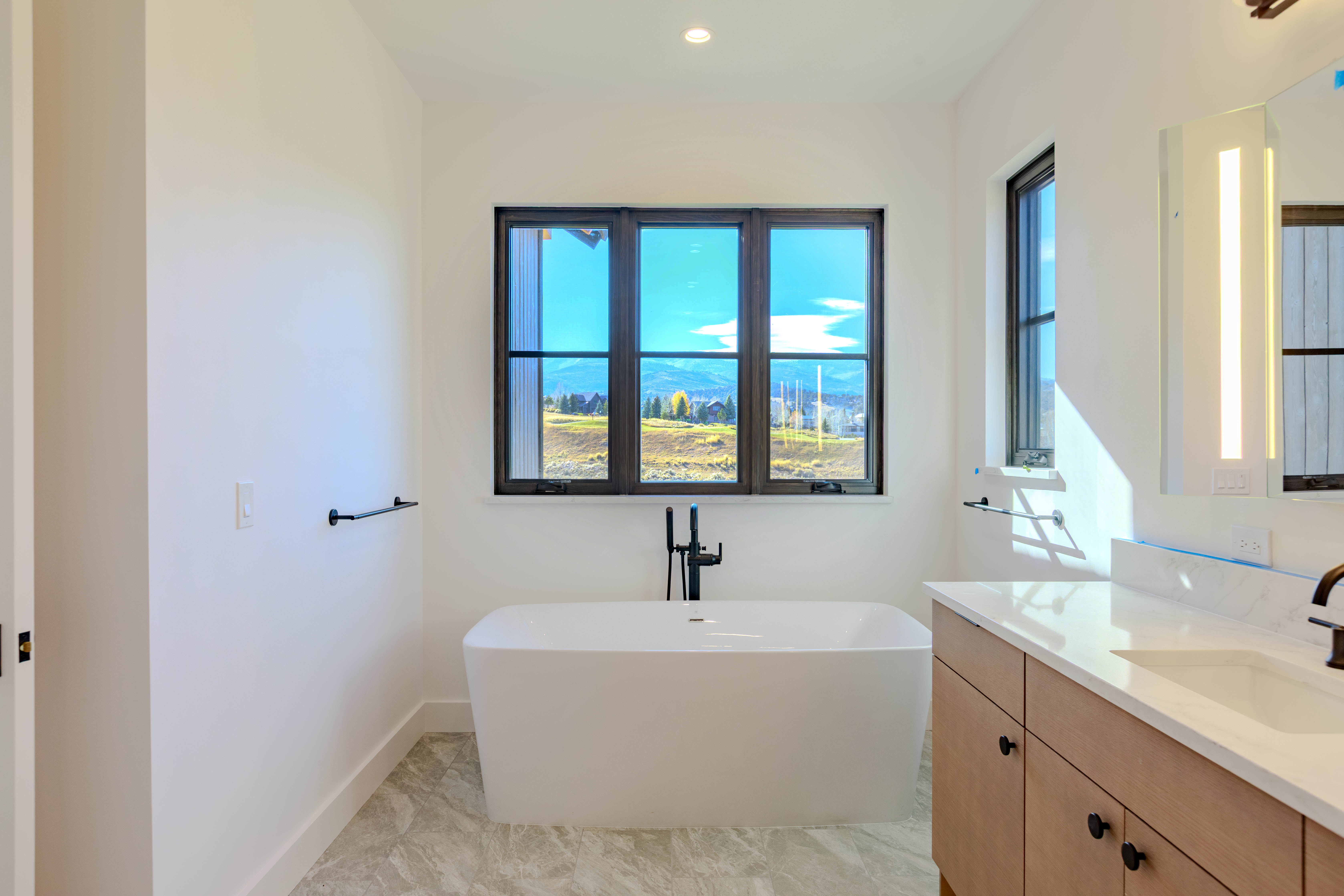 This is a bright and modern primary bathroom featuring a freestanding white bathtub positioned in front of a large window with a scenic view. The bathroom includes a light wood vanity with a white countertop and a sleek, illuminated mirror. The overall aesthetic is clean, luxurious, and designed to maximize natural light and views.