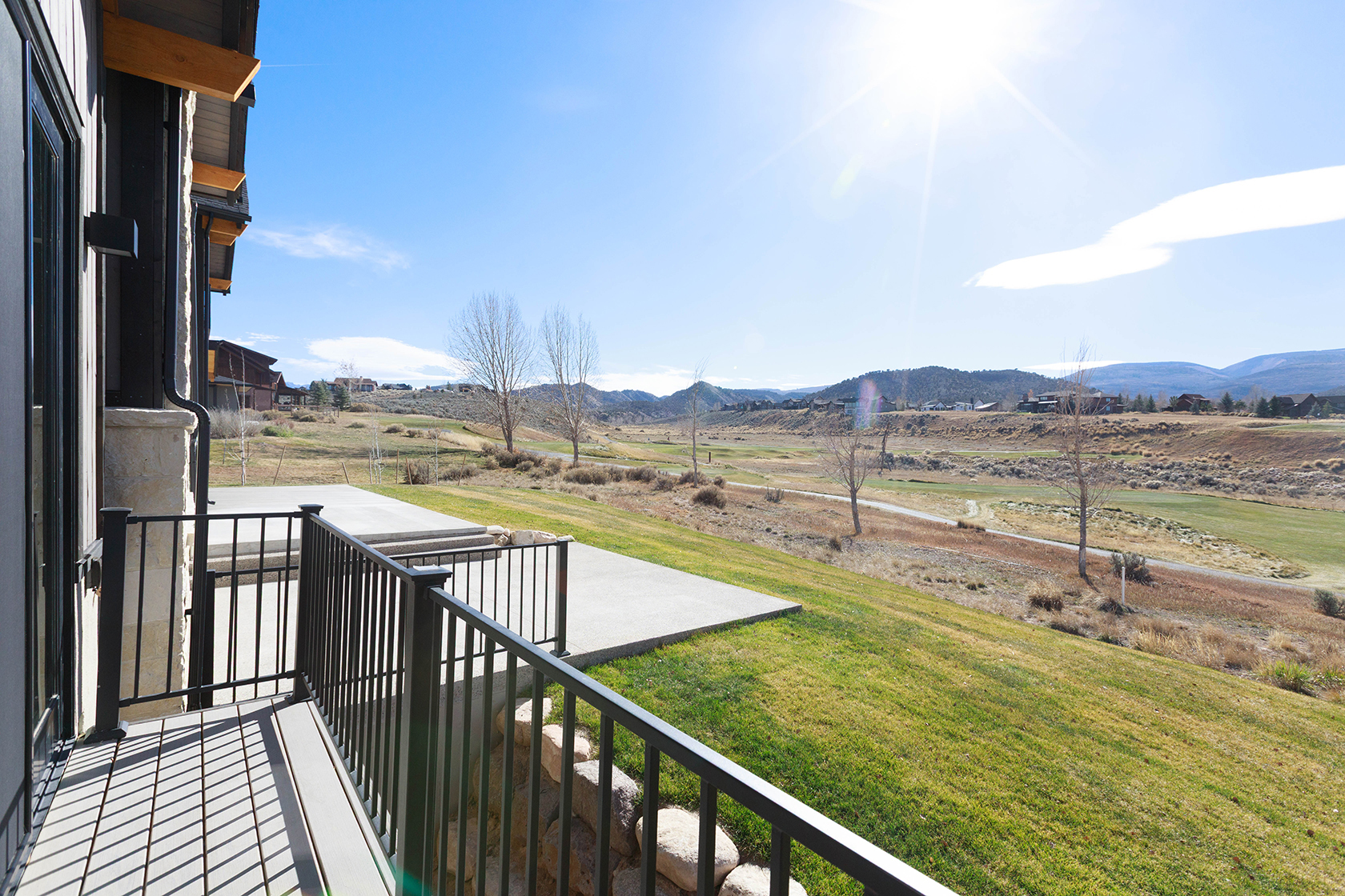 This image showcases a beautiful outdoor patio/deck/balcony area with a black metal railing, offering stunning views of a well-maintained lawn, a golf course, and distant mountains under a clear blue sky. The concrete patio extends from the building, providing an inviting space for relaxation and entertainment. The overall impression is one of luxury and tranquility, highlighting the property's desirable outdoor living space and scenic surroundings.