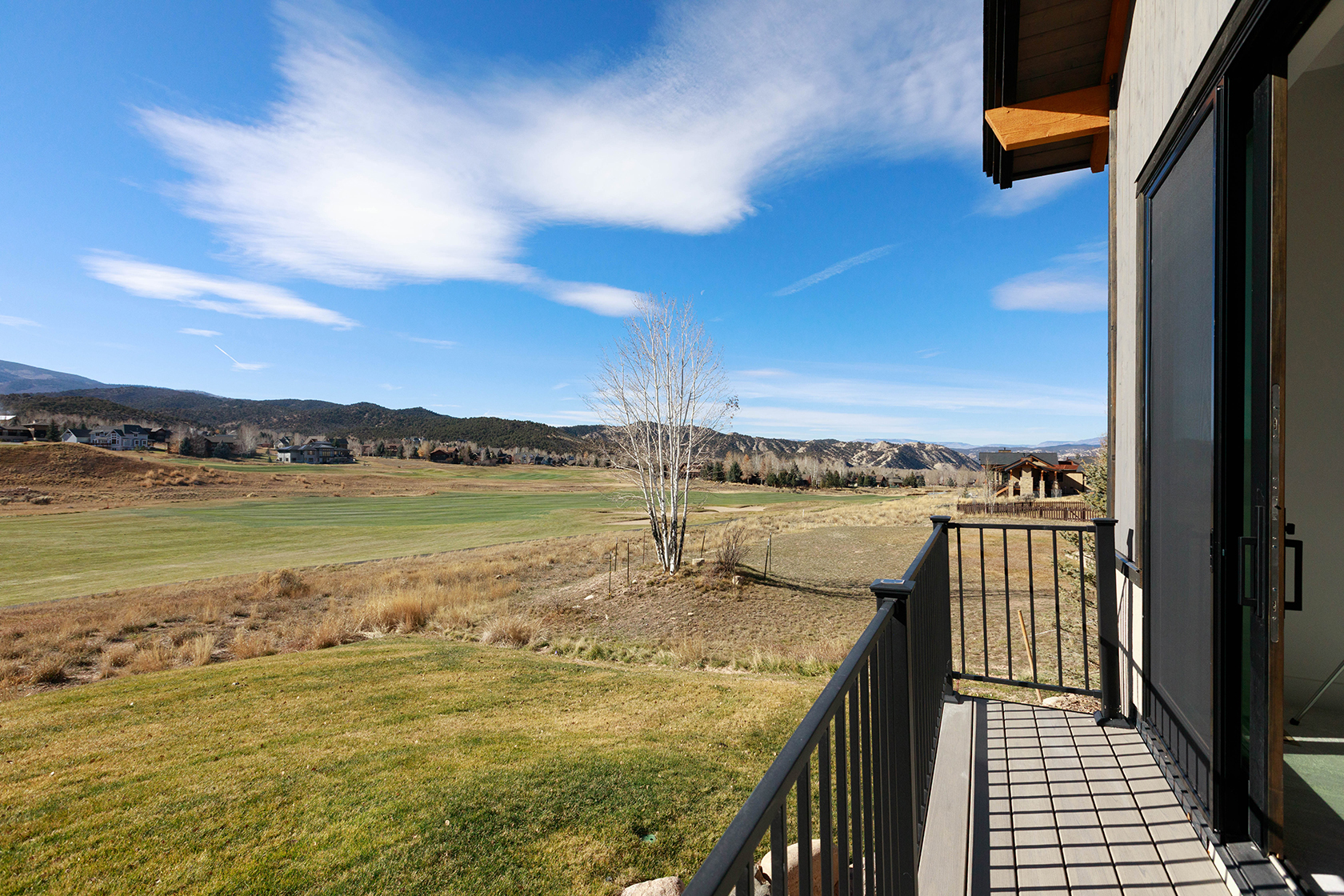 This image showcases a balcony with a view of a sprawling green landscape, likely a golf course or large yard. The balcony features dark metal railings and a composite deck. A sliding glass door provides access to the interior, suggesting a seamless indoor-outdoor living experience. The overall impression is one of tranquility and connection with nature.