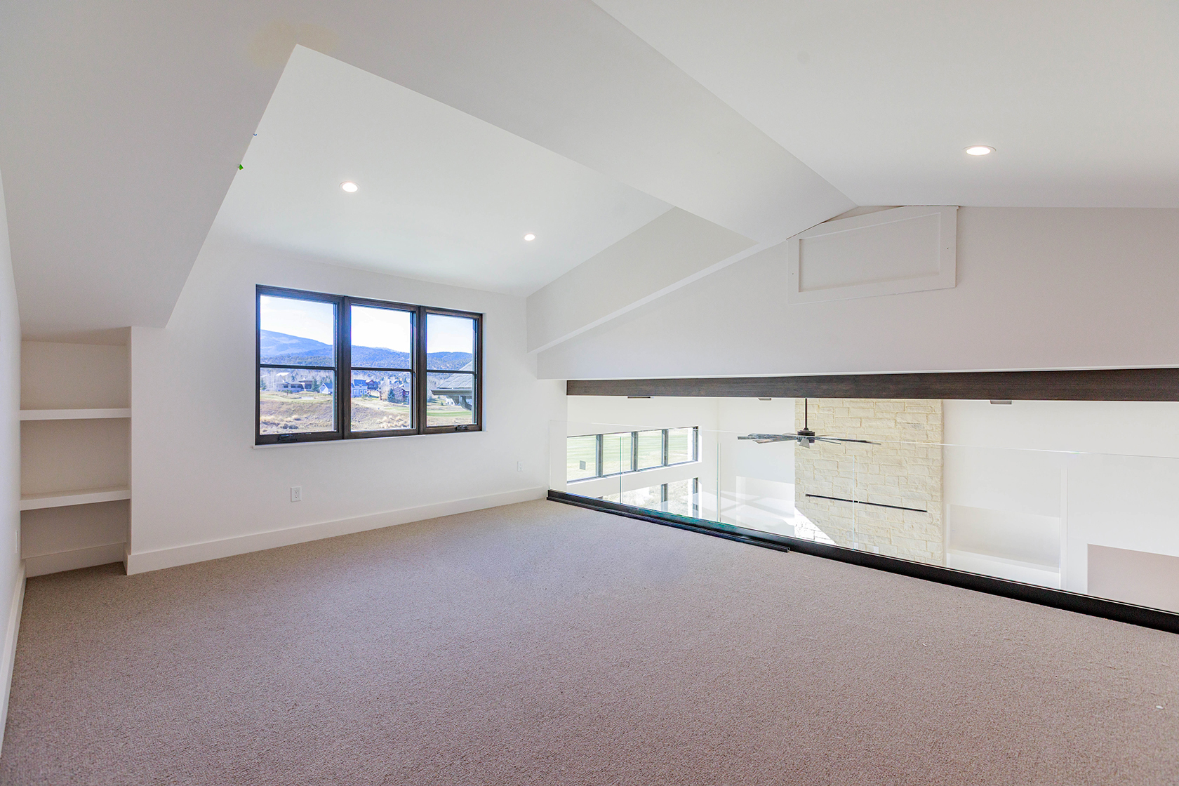 This interior shot showcases a modern hallway or landing area with a view overlooking a lower level. The space features neutral carpet, white walls, and a large window providing natural light and a scenic view. A glass railing provides a contemporary touch, while the open design creates a sense of spaciousness and connection between levels.