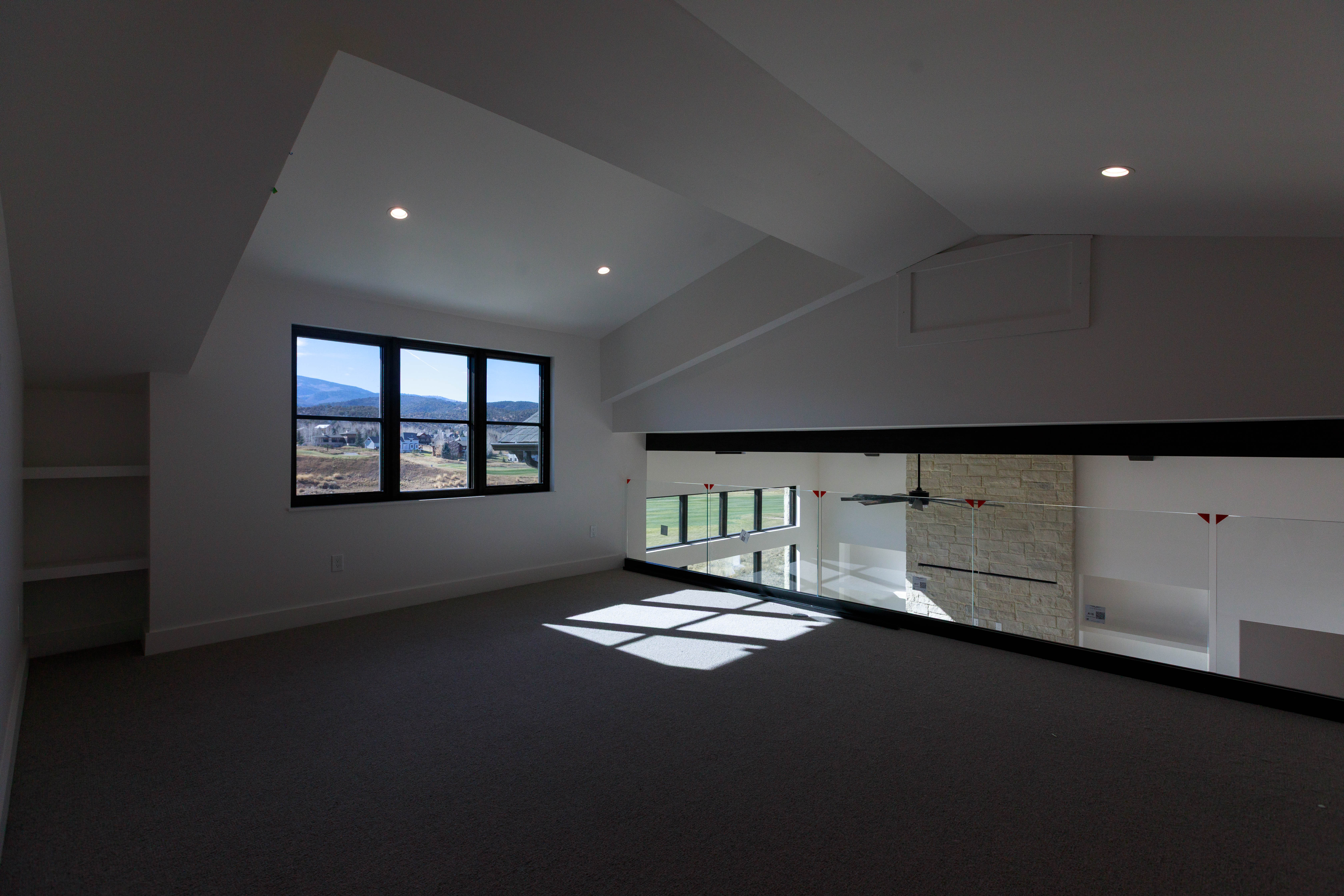 This interior shot showcases a modern hallway or landing area with a view overlooking a lower level. The space features a large window providing natural light, neutral-toned carpeting, and a glass railing system. The design is clean and contemporary, emphasizing open space and natural light.