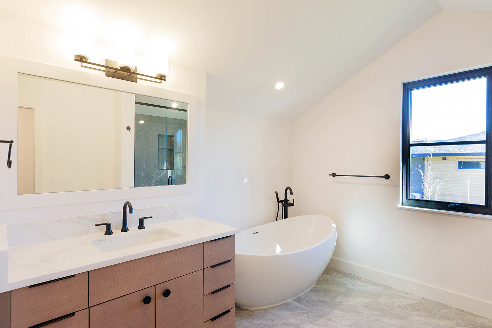 This is a bright and modern primary bathroom featuring a freestanding white bathtub, a wood-finish vanity with a white countertop and black hardware, and a large mirror with integrated lighting. A black framed window provides natural light, and the gray tile flooring adds a touch of elegance. The perspective is a wide shot, capturing the entire space.