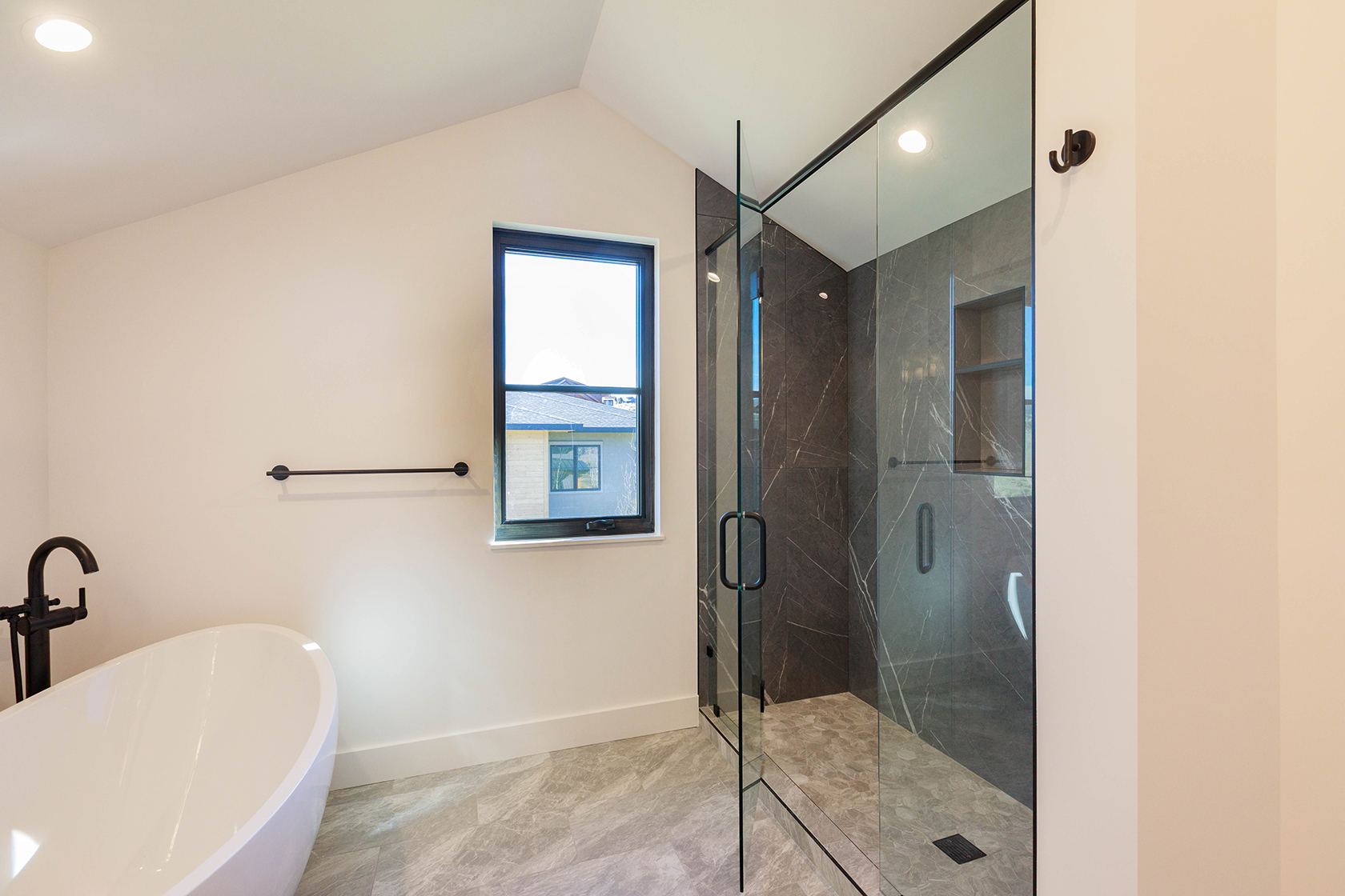 This is a bright and modern primary bathroom featuring a freestanding white bathtub with a black faucet, a sleek glass-enclosed shower with dark marble-like tiling, and a black-framed window. The walls are painted in a neutral tone, and the flooring is a light gray marble, creating a clean and luxurious aesthetic. The perspective is from the doorway, showcasing the entire bathroom.