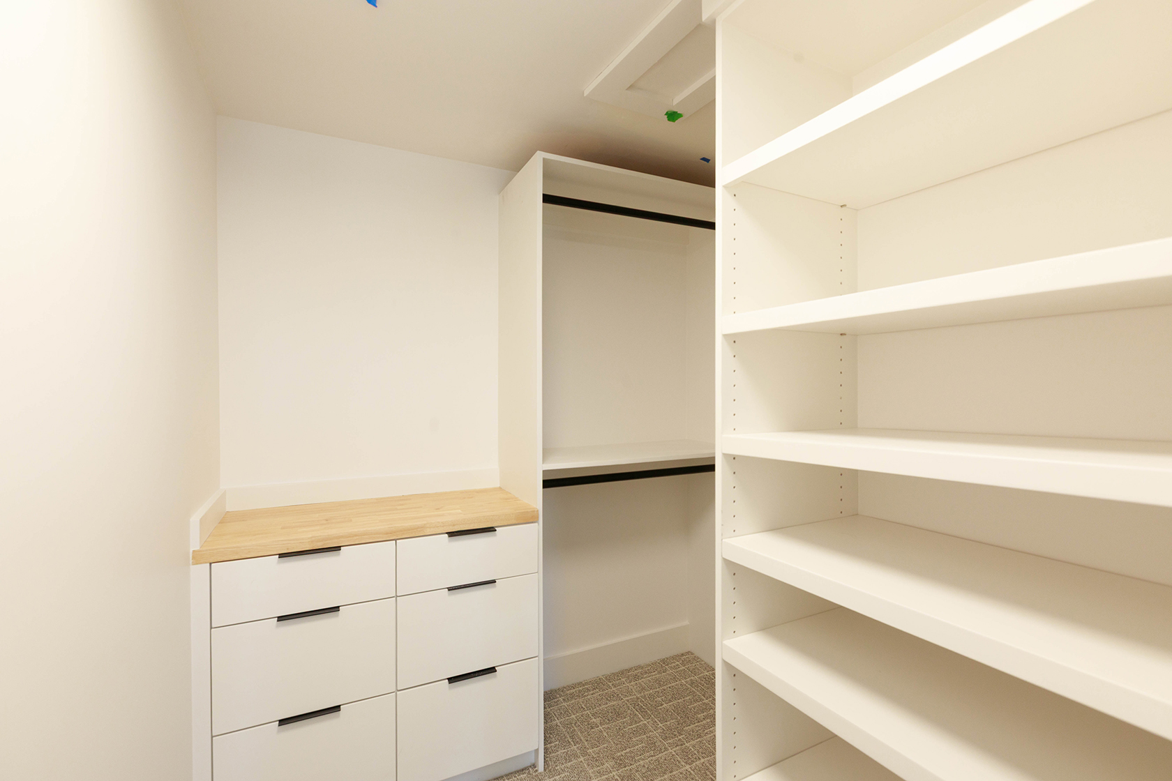 This is a well-organized walk-in closet featuring white shelving and drawers with modern black handles. The closet includes a section for hanging clothes and open shelving for folded items or storage. The overall impression is clean, bright, and functional, maximizing storage space.
