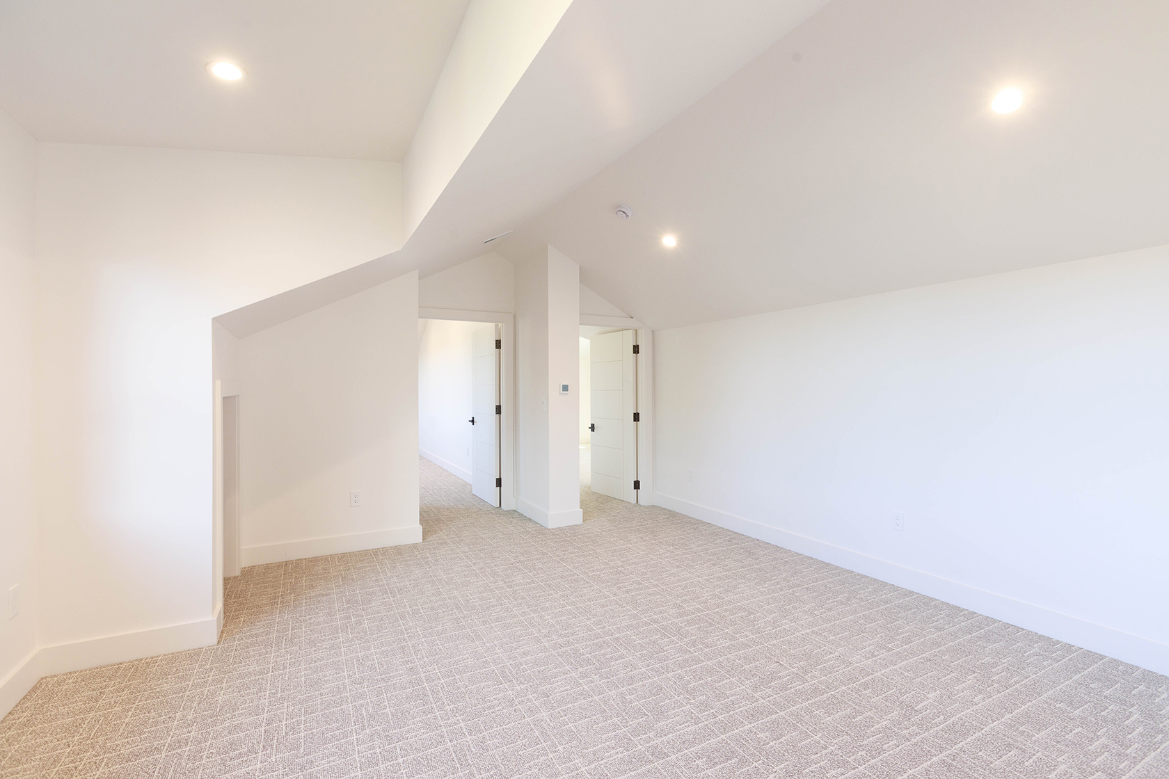This is a bright and spacious bedroom with neutral-toned carpet and white walls, creating a clean and modern aesthetic. The room features recessed lighting and two doorways, suggesting access to other areas of the house. The overall impression is one of simplicity and potential for customization.