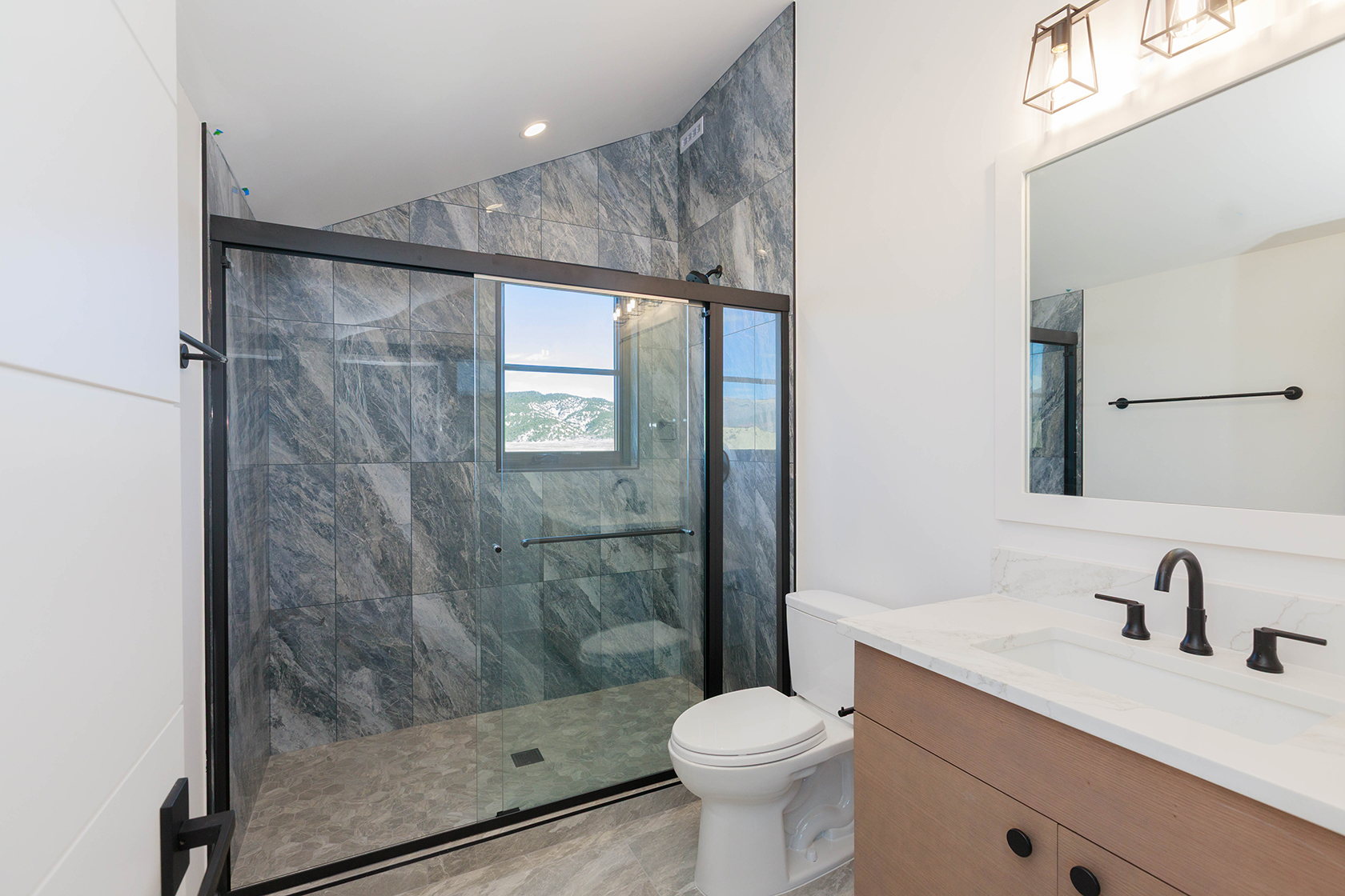 This is a modern bathroom featuring a walk-in shower with gray marble-style tiling and a black framed glass door. The vanity has a light wood cabinet, white countertop, and black fixtures. A large mirror hangs above the sink, and the toilet is white. The perspective is from the doorway, showcasing the entire bathroom.