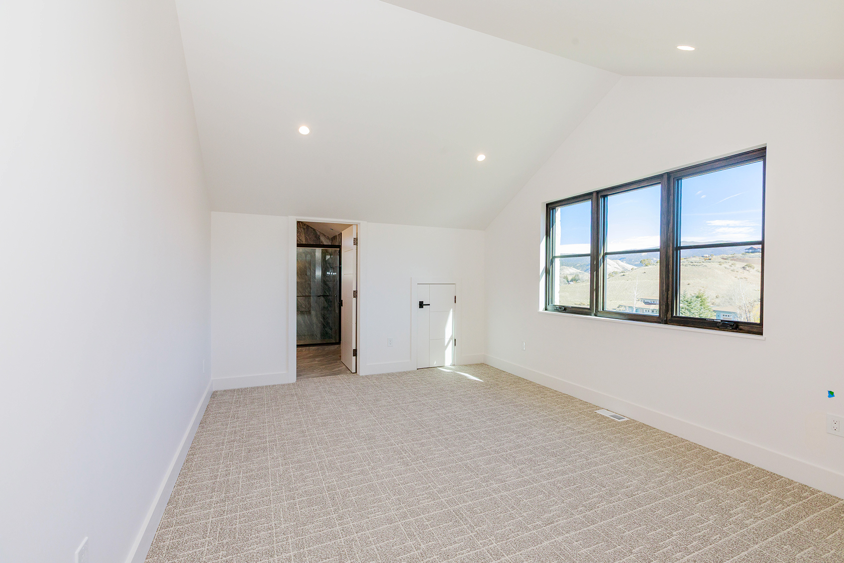 This is a bright and spacious primary bedroom featuring neutral-toned carpet and white walls, creating a serene atmosphere. The room has a vaulted ceiling and a large window offering natural light and views of the landscape. There is an ensuite bathroom and a closet, adding to the functionality of the space.