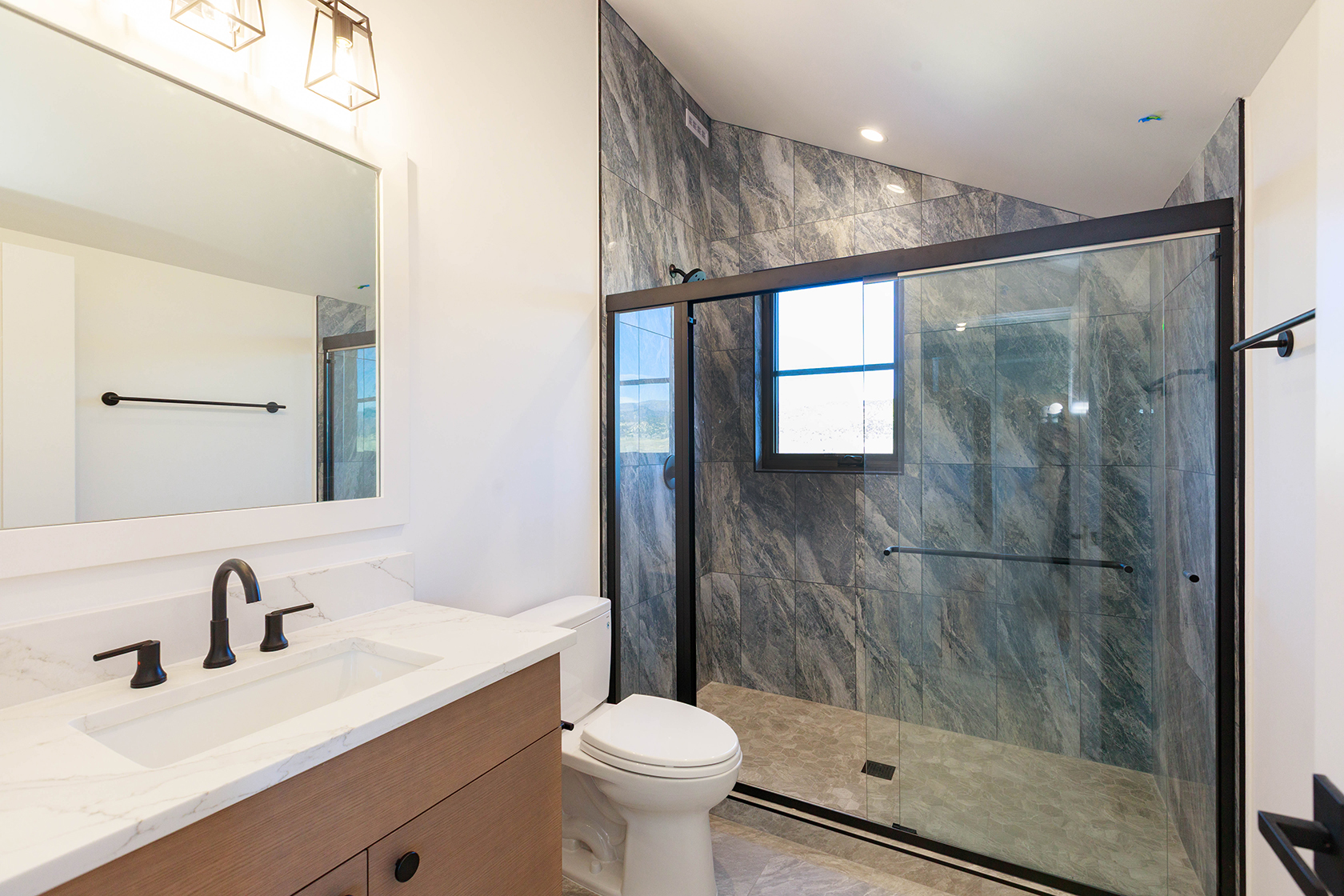 This is a well-lit primary bathroom featuring a modern vanity with a white countertop and wood-grain cabinets, complemented by matte black fixtures. A spacious walk-in shower with gray marble-style tiling and a glass enclosure is visible. The overall design is sleek and contemporary, creating a luxurious and functional space.
