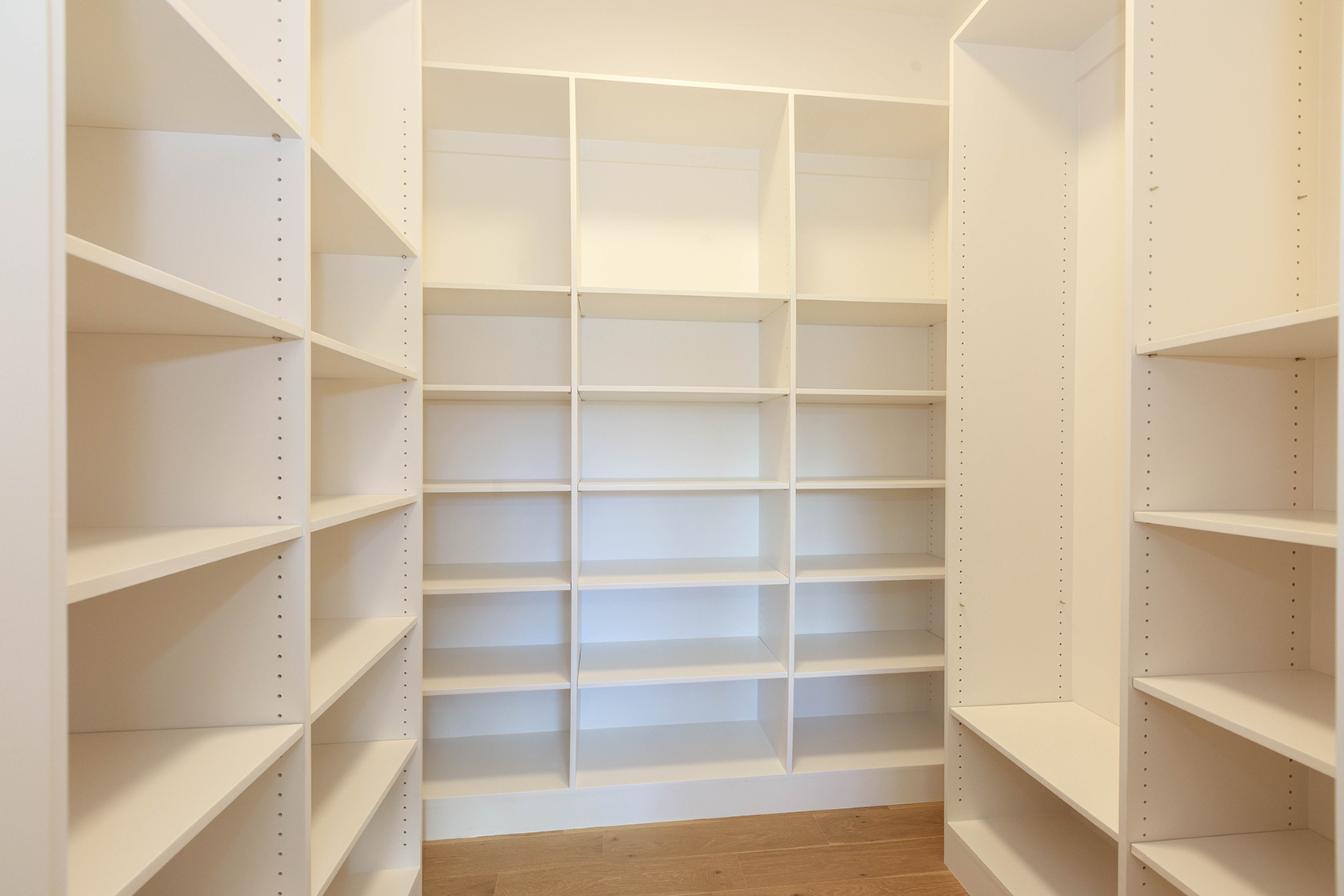 This is a well-organized walk-in closet featuring custom shelving and storage solutions. The closet is painted in a neutral color, providing a clean and bright aesthetic. The shelves are empty, ready to be filled with clothes and accessories, and the wooden floor adds a touch of warmth.