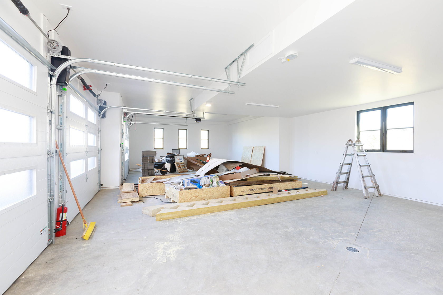 This is a spacious garage with a white garage door partially open, revealing a well-lit interior. The garage features a concrete floor, white walls, and a high ceiling with overhead lighting. Construction materials and a ladder are present, suggesting the space is either under renovation or used for storage.