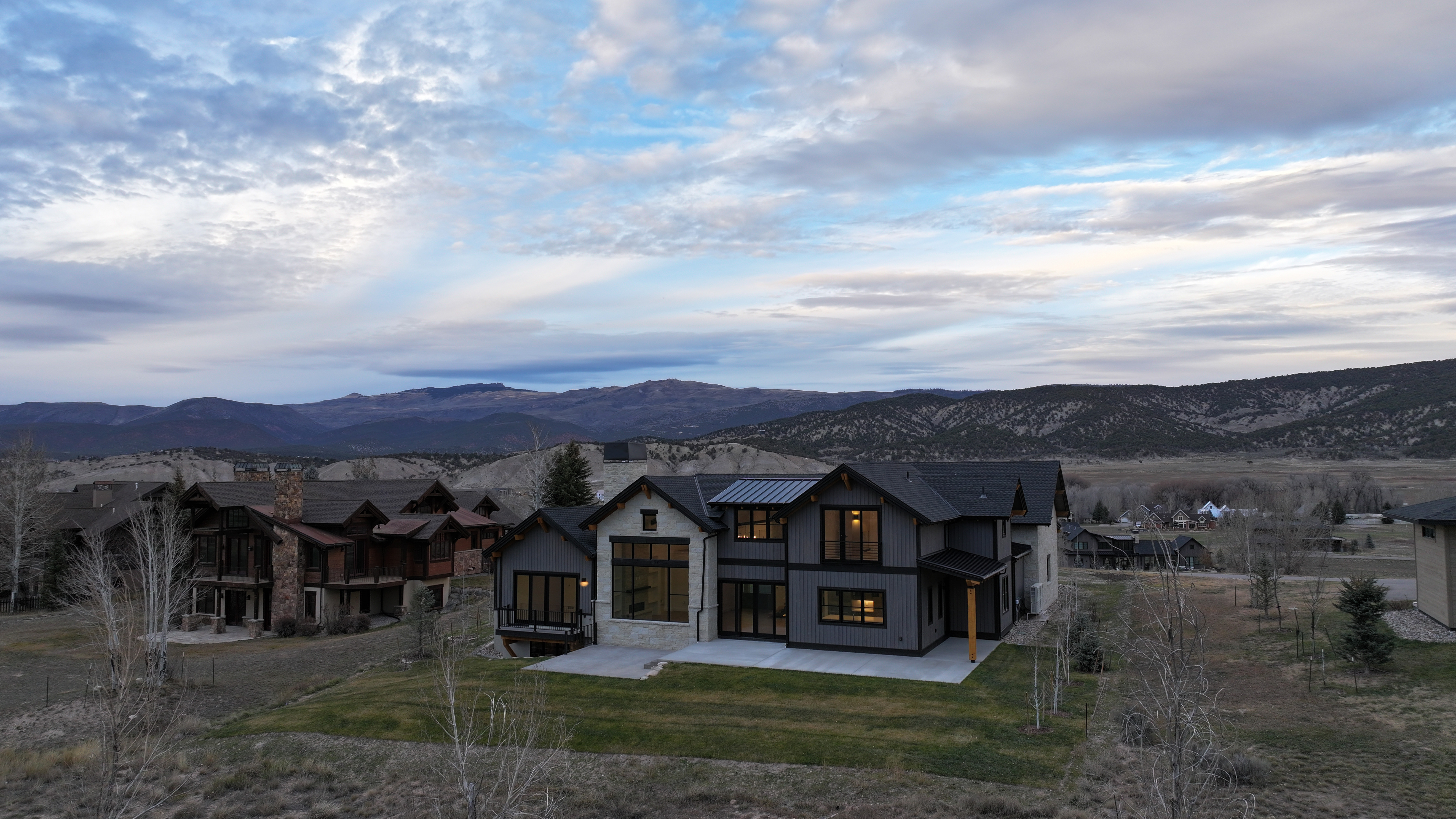 This aerial view showcases a modern, two-story home with a combination of stone and dark siding, complemented by large windows and a well-maintained lawn. The house is situated in a scenic location with rolling hills and mountains in the background, creating a sense of privacy and natural beauty. The surrounding landscape includes other residential properties, blending the home into a community setting.