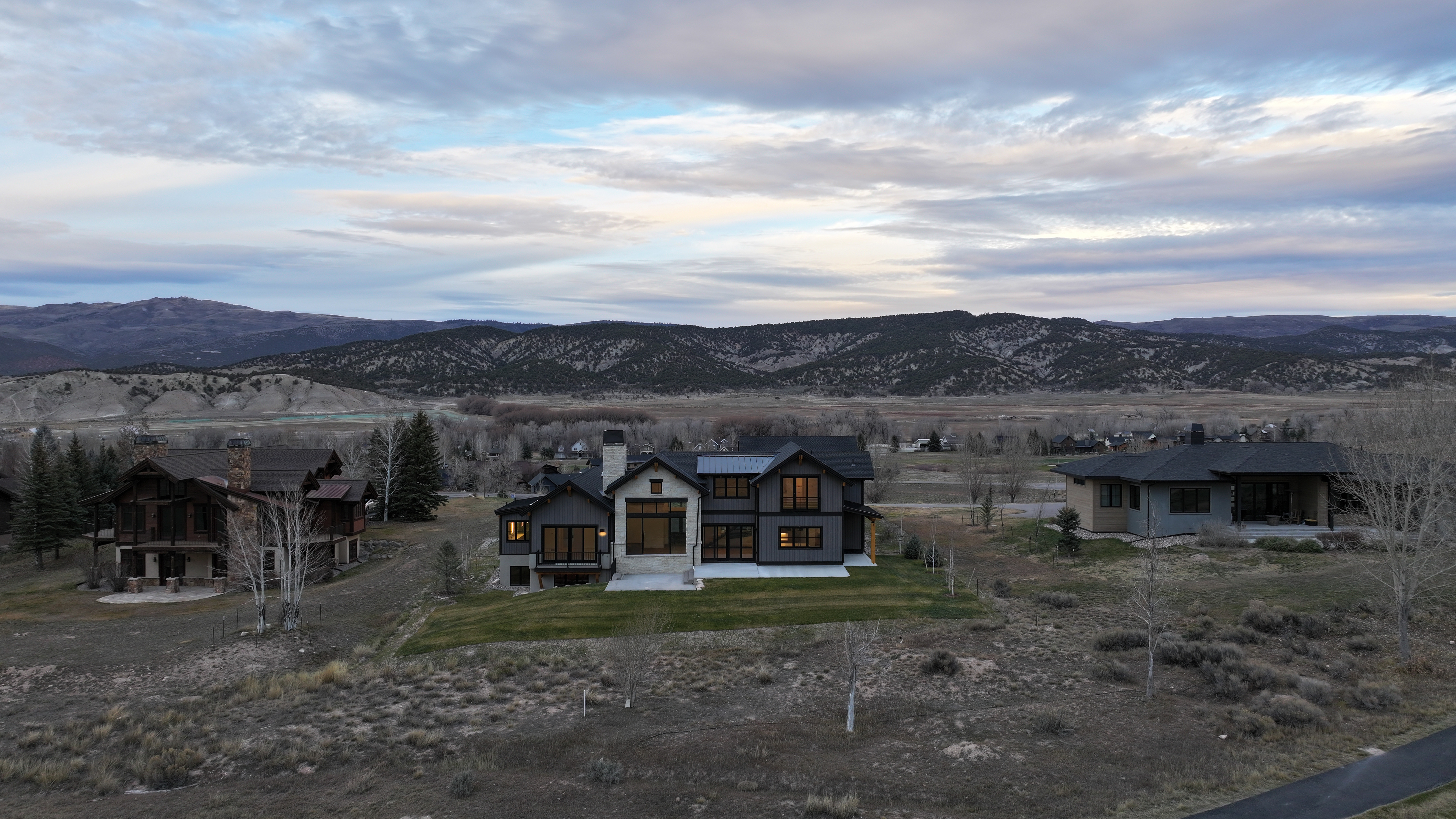 This aerial view showcases a modern home with a dark exterior and light stone accents, situated on a well-manicured lawn. The property is surrounded by a natural landscape with rolling hills and sparse vegetation, creating a sense of privacy and seclusion. Other houses are visible in the distance, and the sky is filled with clouds, adding depth and texture to the scene.
