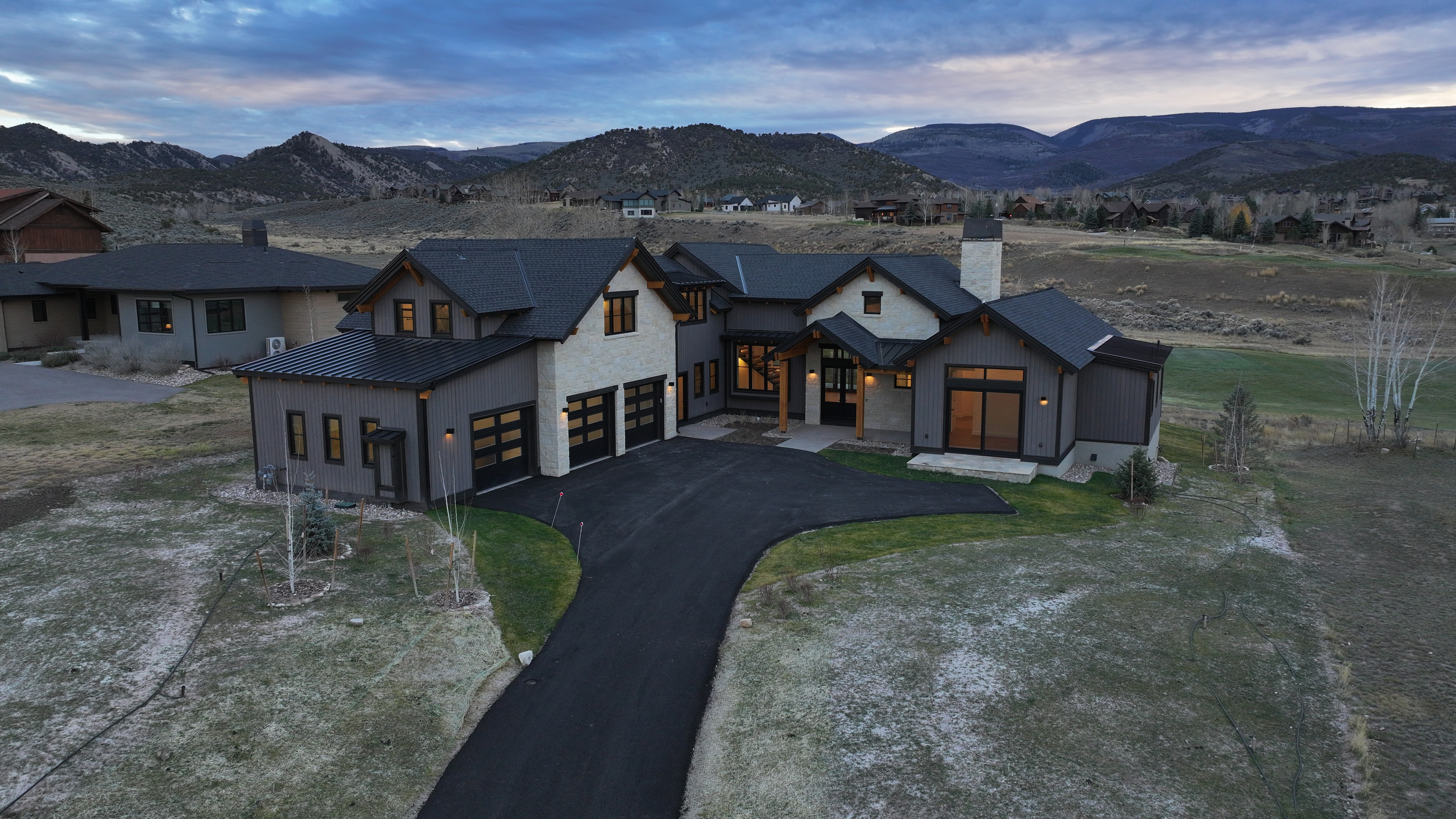 This aerial view showcases a modern luxury home with a dark roof, gray siding, and stone accents. The property features a paved driveway, well-maintained landscaping, and is situated in a scenic mountain setting. The home's design emphasizes clean lines and a sophisticated aesthetic.
