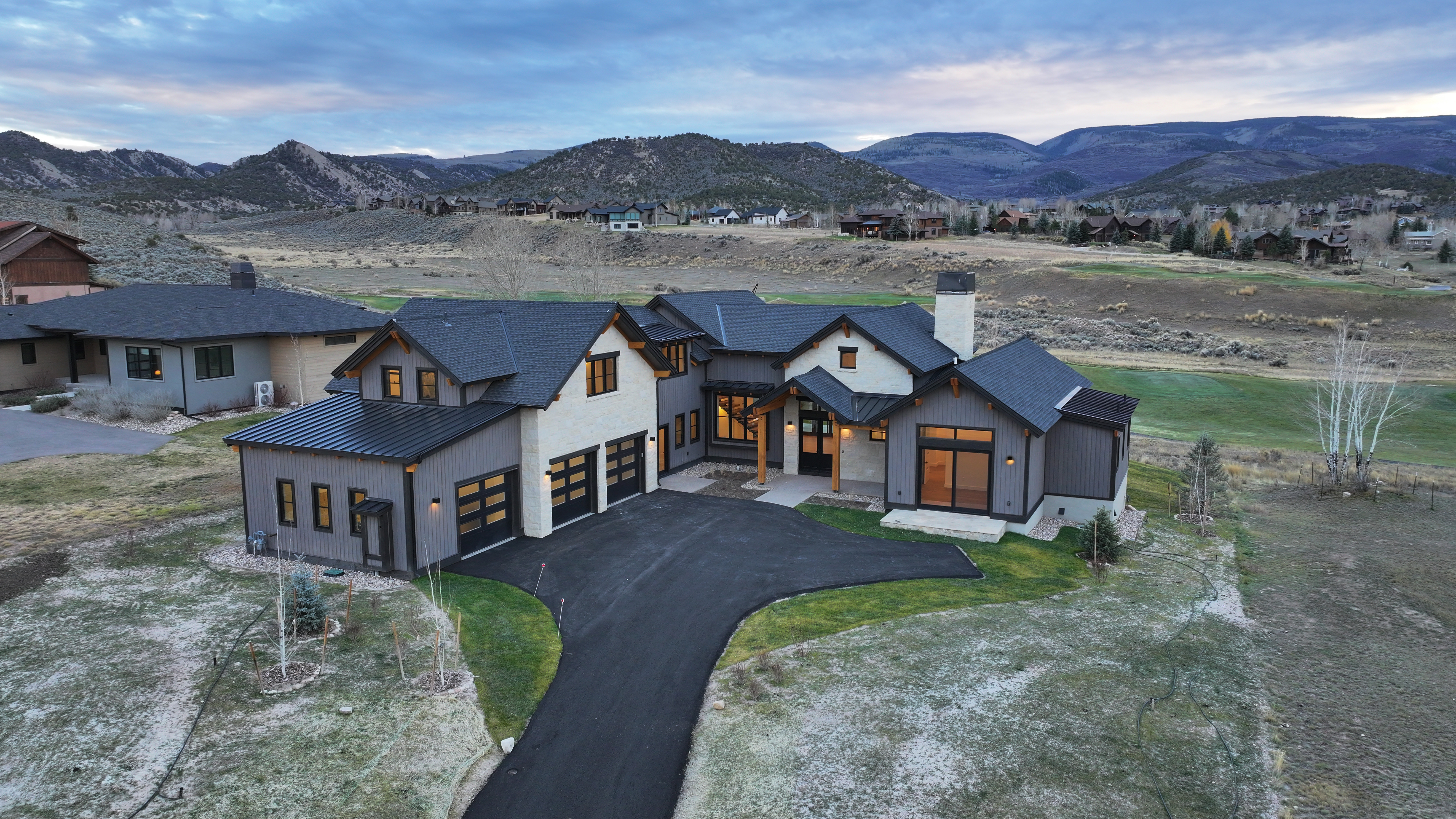 This aerial view showcases a luxurious modern home nestled in a scenic, mountainous landscape. The house features a combination of gray siding and stone accents, a dark roof, and a well-manicured lawn that blends seamlessly with the natural surroundings. A winding driveway leads to the attached garage, enhancing the property's curb appeal and highlighting its spacious setting.