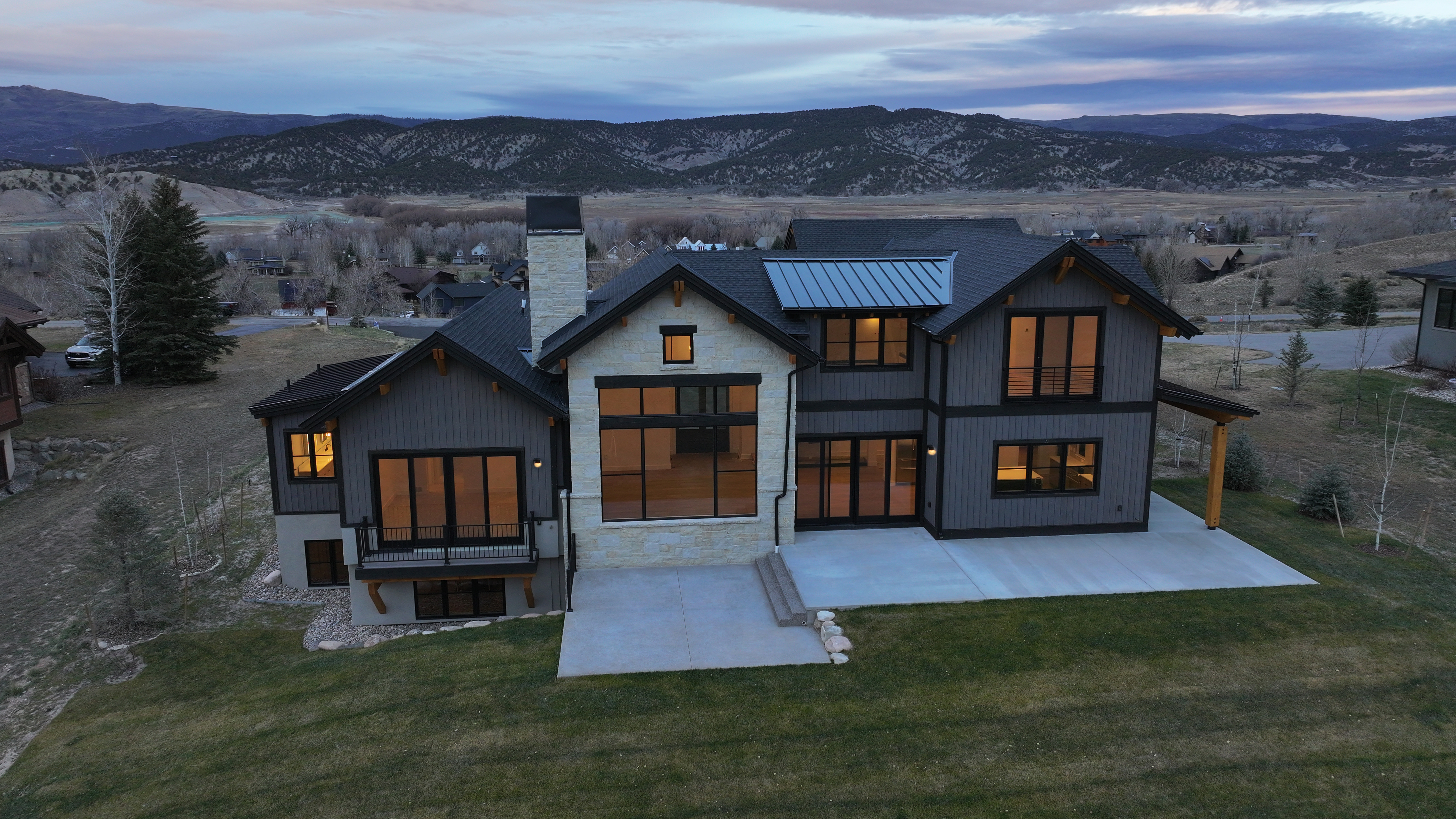 This is an aerial rear view of a modern, two-story home with a combination of stone and dark gray siding. The house features large windows and doors leading to a concrete patio and a well-maintained lawn. In the background, there are rolling hills and a partially cloudy sky, adding to the property's scenic appeal.