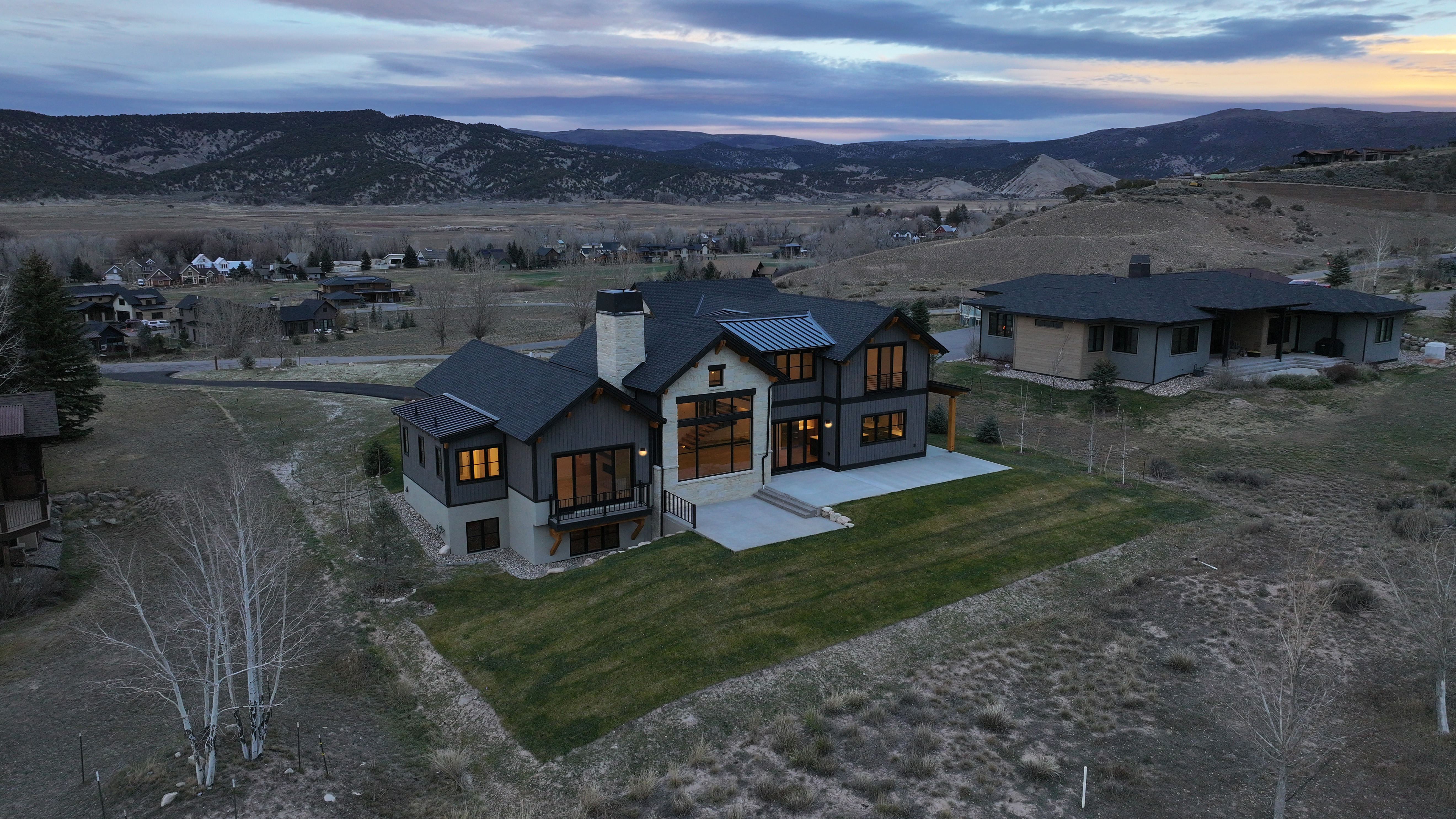 This aerial view showcases a modern, two-story home with a dark gray exterior, light stone accents, and a well-manicured lawn. The property is situated on a hillside with mountain views in the background, offering a sense of privacy and luxury. A neighboring house is visible to the right, and the overall setting is serene and upscale.