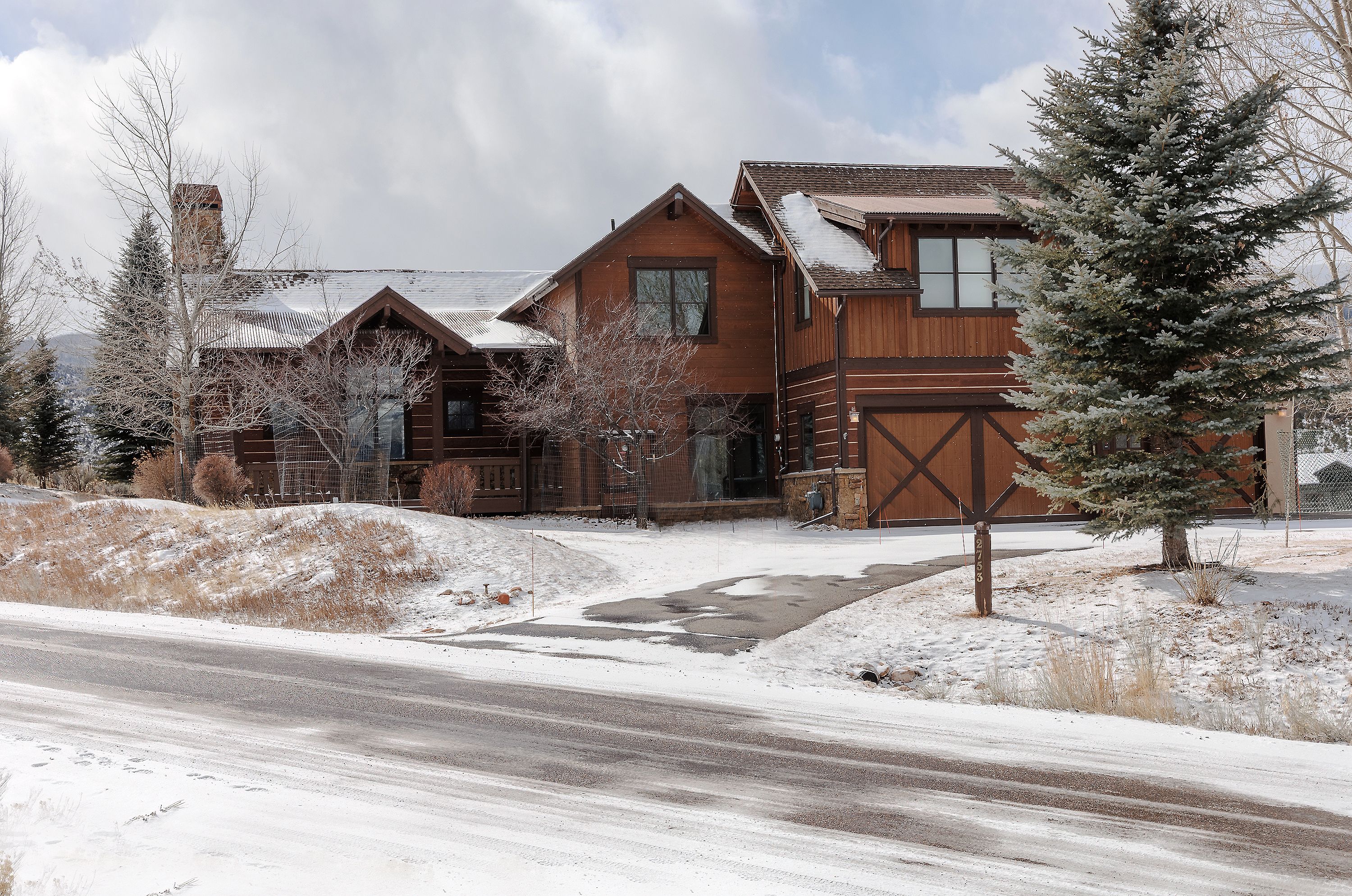 This charming mountain-style home features a rustic wood-paneled exterior with a prominent garage and a stone-accented foundation. The property is set against a snowy landscape with a mature evergreen tree in the foreground, creating a cozy and inviting winter atmosphere. The perspective captures the full front facade from the street, highlighting the architectural details and the home's integration into its natural surroundings.
