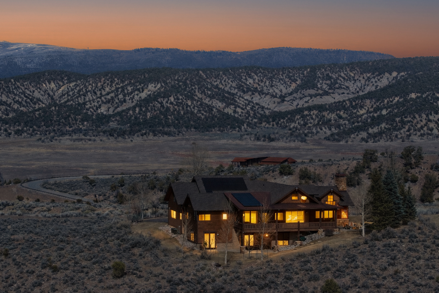 This stunning aerial view captures a rustic, multi-level wooden lodge nestled within a vast, rugged landscape during a serene sunset. The home features warm, inviting interior lighting, a spacious deck, and solar panels on the roof, all set against a backdrop of rolling hills and distant snow-capped mountains. The composition emphasizes the property's secluded, tranquil setting and architectural integration with the natural environment.