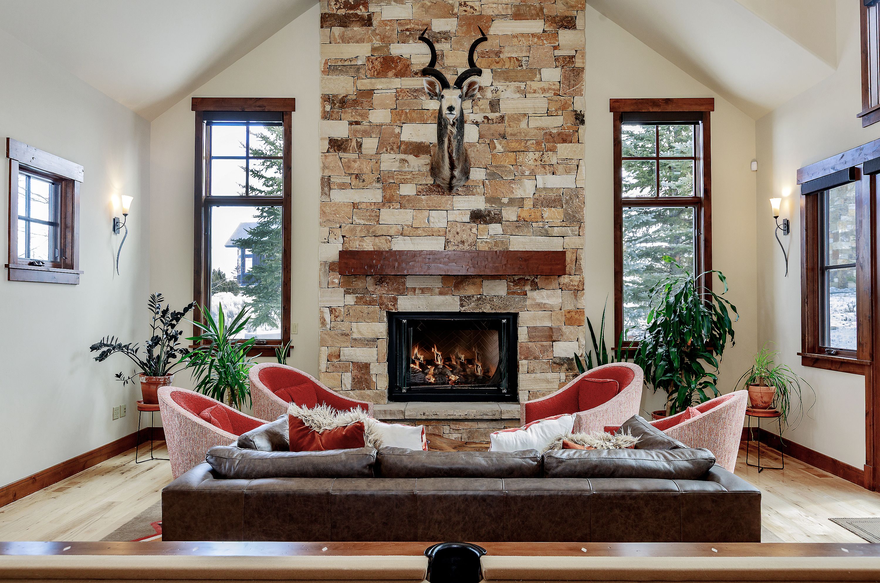 This inviting living room features a grand, floor-to-ceiling stone fireplace as its focal point, adorned with a mounted animal trophy and a rustic wooden mantel. The space is furnished with a plush, dark leather sofa and four vibrant red accent chairs, creating a cozy and symmetrical seating arrangement. Large windows on either side of the fireplace flood the room with natural light, highlighting the warm wood flooring and vaulted ceilings, perfect for a mountain-style retreat.