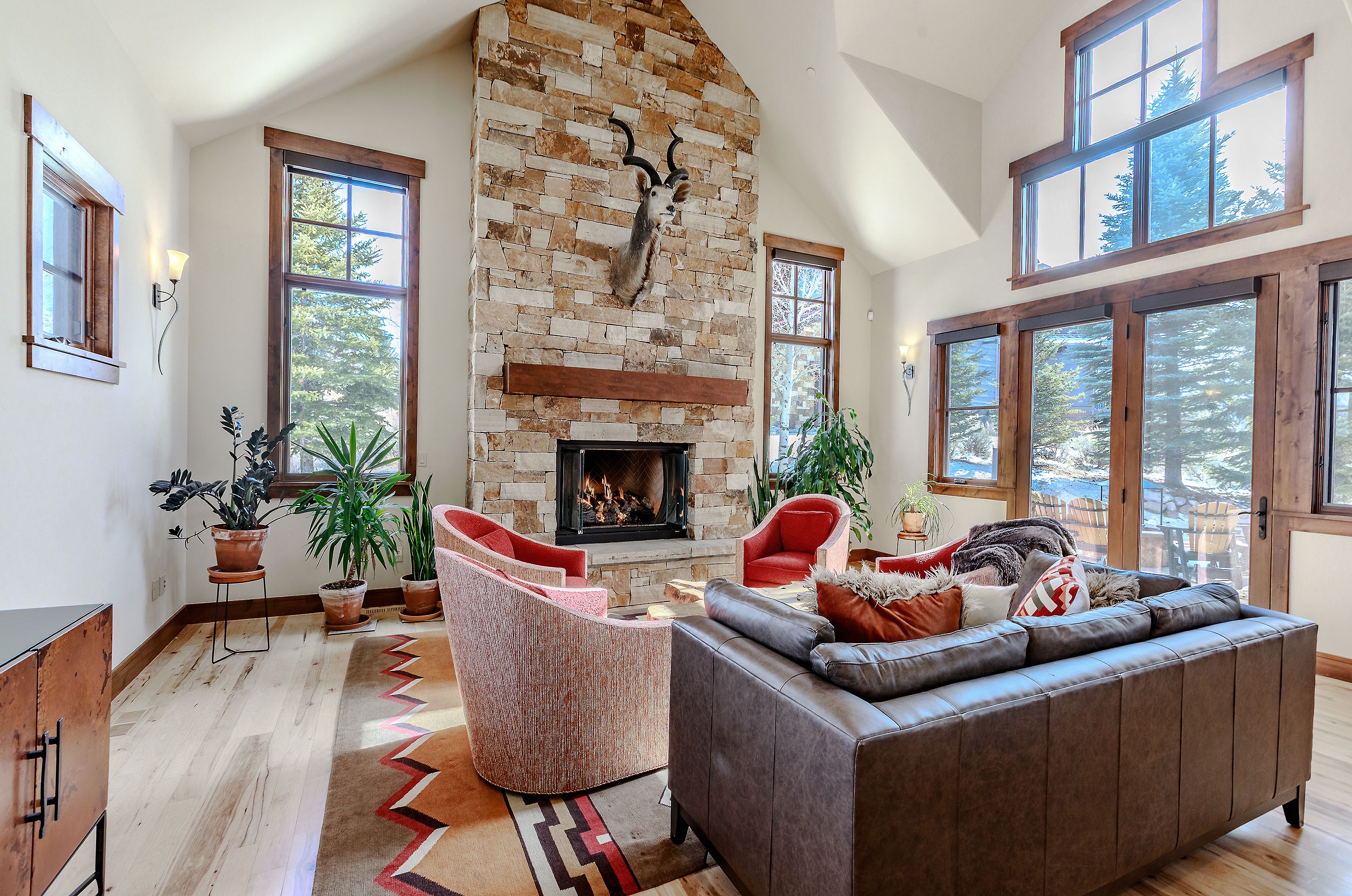 This spacious, high-ceilinged living room features a striking floor-to-ceiling stone fireplace as its focal point, adorned with a mounted animal trophy. The room is furnished with a comfortable leather sofa and plush armchairs arranged on a patterned area rug, creating a cozy, rustic-chic atmosphere. Large windows and glass doors flood the space with natural light, offering scenic views of the surrounding trees and landscape.