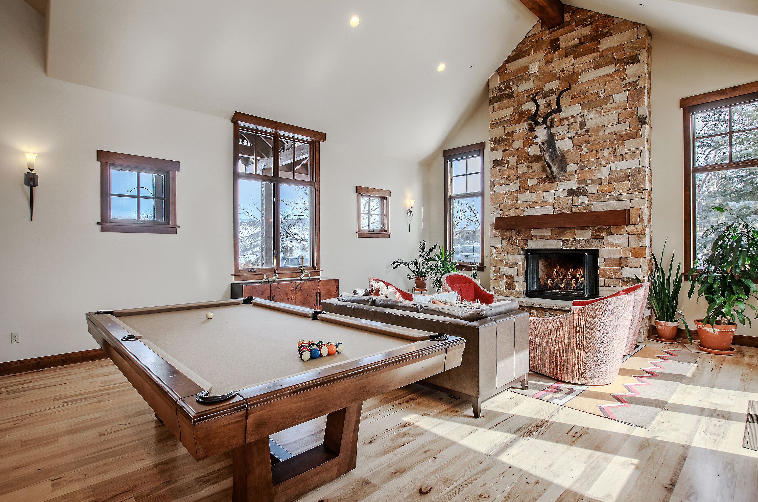 This spacious, open-concept living area features a rustic aesthetic with a vaulted ceiling, light hardwood floors, and a prominent floor-to-ceiling stone fireplace adorned with a mounted animal head. The room is furnished with a pool table in the foreground, a comfortable leather sofa, and two plush armchairs, creating a cozy yet sophisticated atmosphere. Large windows allow for abundant natural light and provide scenic views, making it an ideal space for both entertainment and relaxation.