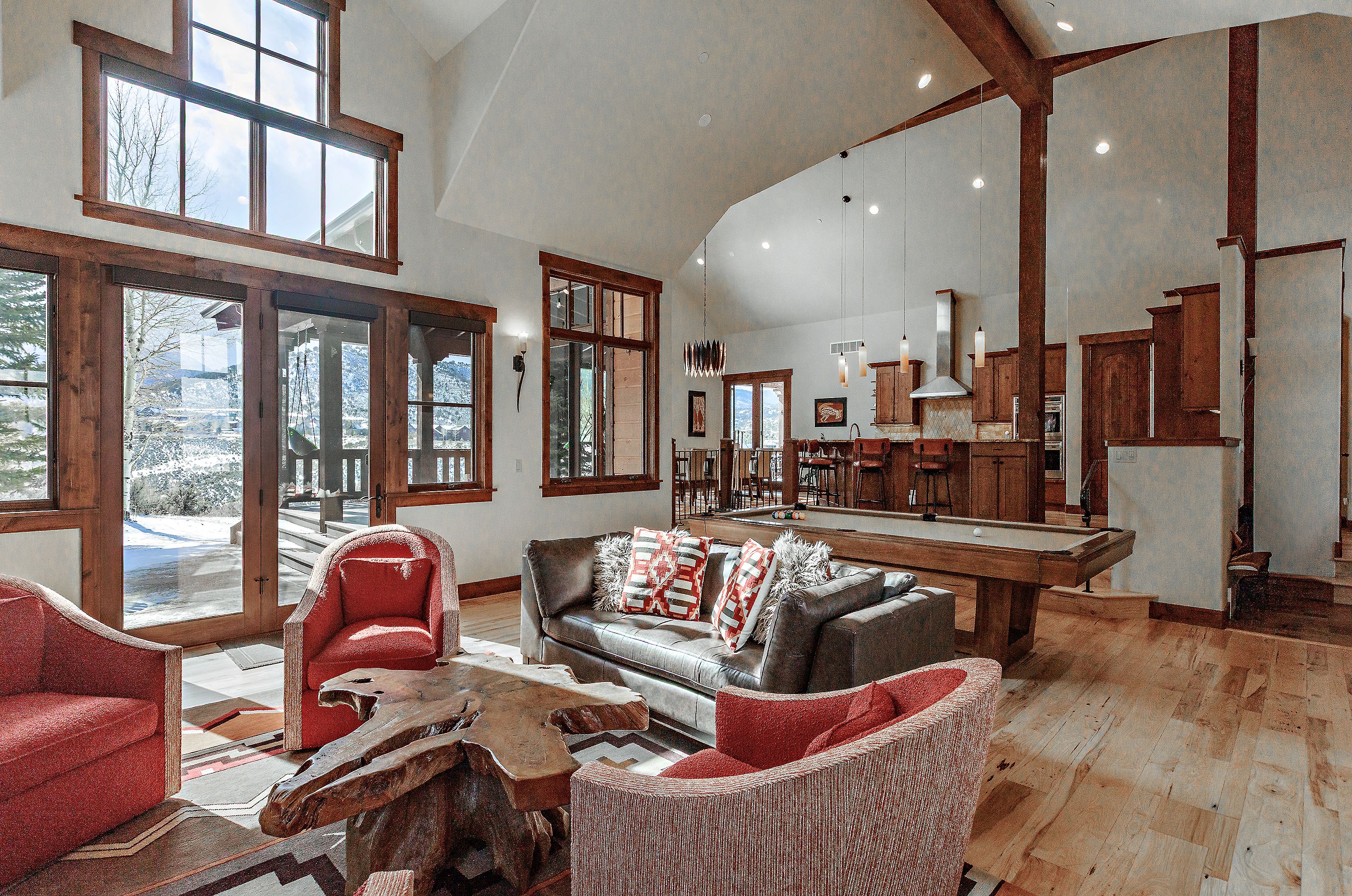 This expansive, open-concept living area features a rustic-modern aesthetic with vaulted ceilings, exposed timber beams, and warm hardwood flooring. The space is anchored by a unique, organic-shaped wood coffee table surrounded by plush red armchairs and a leather sofa, while a pool table sits in the background near the kitchen. Large windows and glass doors provide abundant natural light and scenic views of a snowy landscape, creating a cozy yet grand atmosphere.