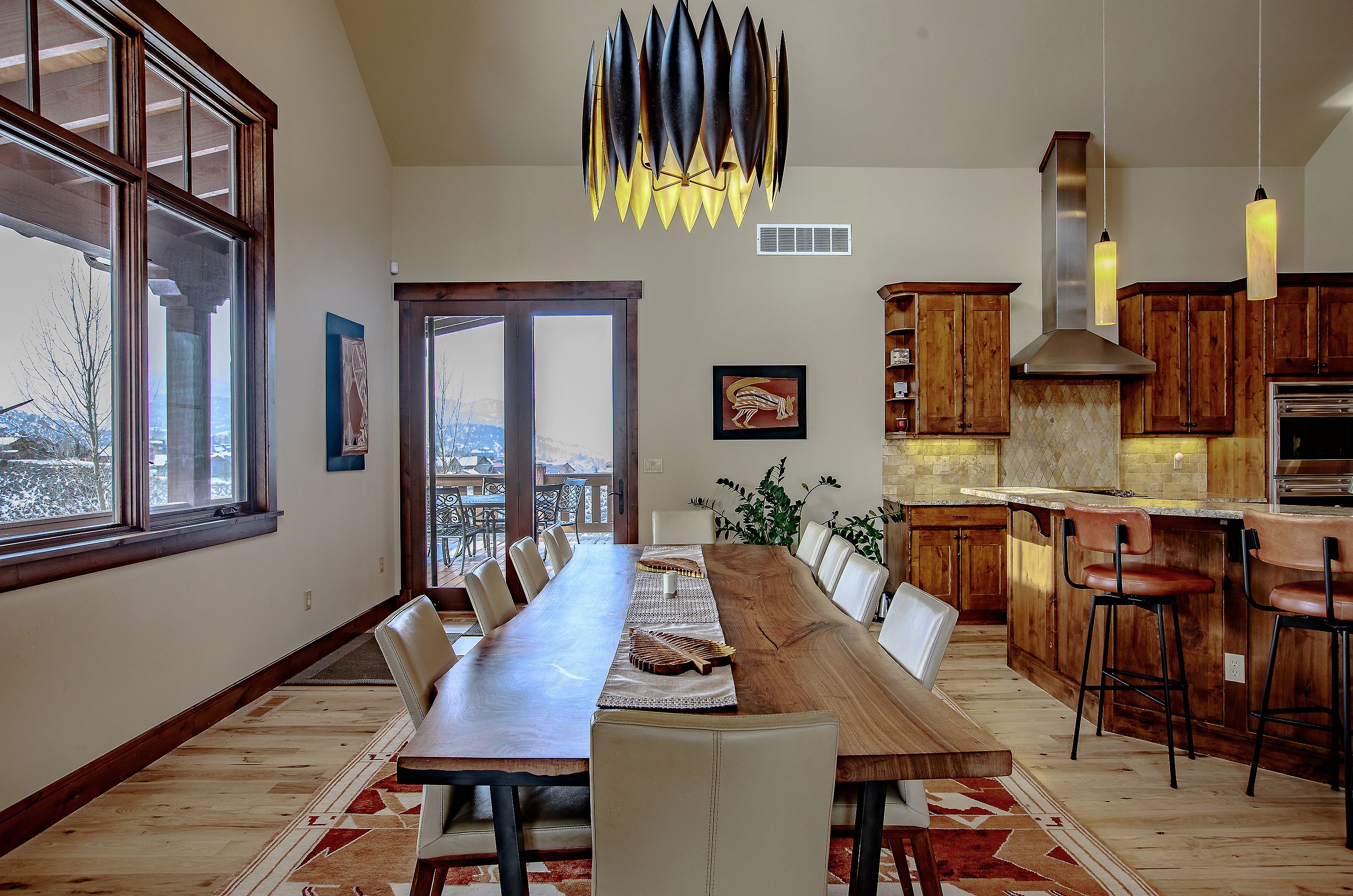 This elegant dining area features a massive, live-edge wood table as its centerpiece, surrounded by modern, cream-colored upholstered chairs. The space is anchored by a striking, contemporary chandelier and a patterned area rug, while an open-concept layout seamlessly connects it to a rustic-style kitchen with wooden cabinetry and a breakfast bar. Large windows and glass doors provide abundant natural light and a scenic view of the snow-covered mountains outside, creating a warm and inviting atmosphere.