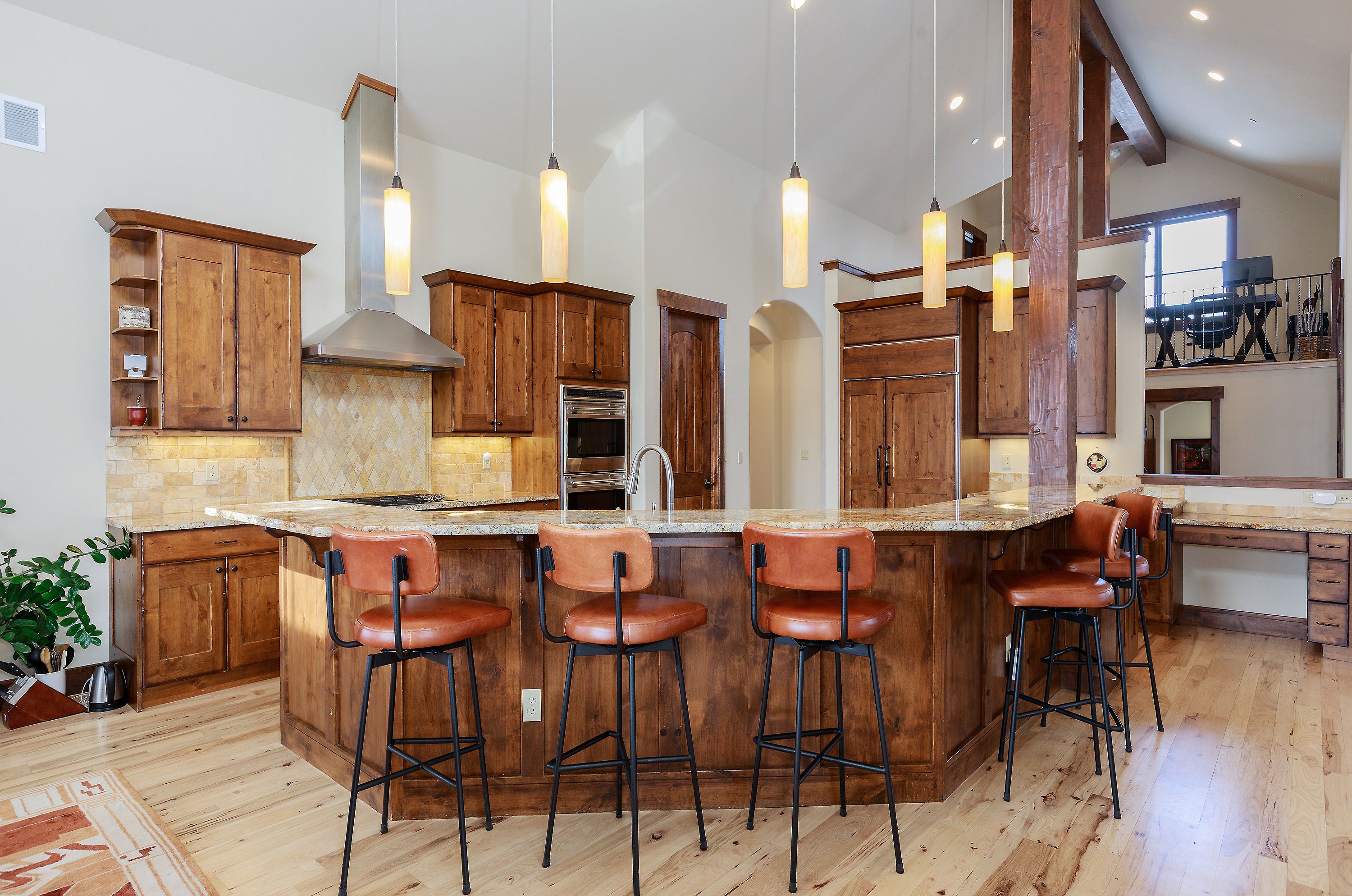 This spacious, rustic-style kitchen features warm wood cabinetry, a large granite-topped island with four leather bar stools, and high vaulted ceilings. The space is illuminated by a series of elegant pendant lights and includes stainless steel appliances, a built-in oven, and a tiled backsplash. The open-concept design flows seamlessly into a lofted area, creating an inviting and functional atmosphere perfect for entertaining.