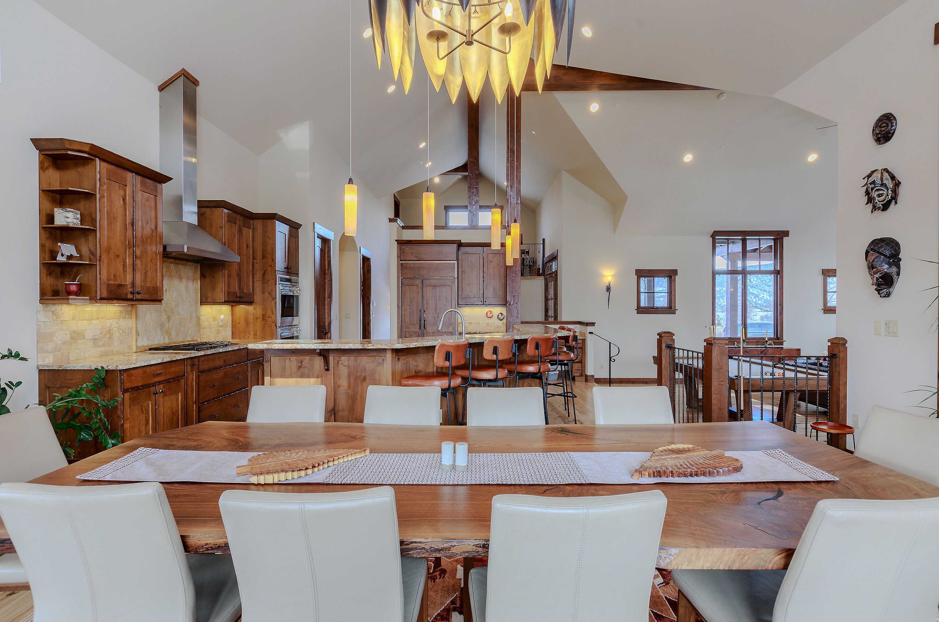 This expansive open-concept space features a large, natural-edge wood dining table as the centerpiece, seamlessly flowing into a gourmet kitchen with warm wood cabinetry and a breakfast bar. The room is defined by soaring vaulted ceilings, exposed timber beams, and a striking, modern chandelier that anchors the dining area. The warm, rustic-modern aesthetic is complemented by high-end finishes and a view into the adjacent living area, creating an inviting and luxurious atmosphere.