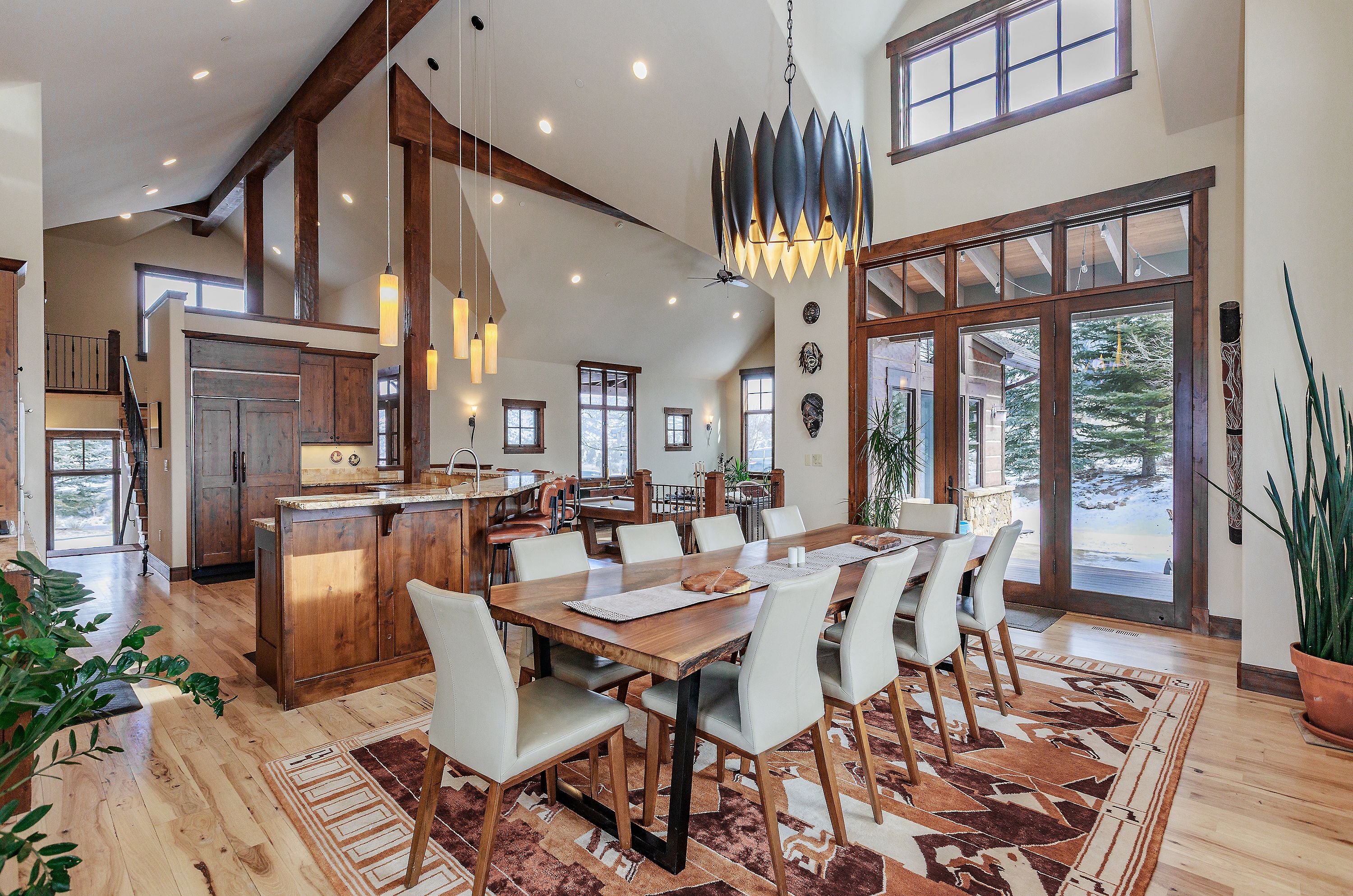 This spacious, open-concept dining area features a large live-edge wooden table surrounded by modern, light-colored chairs, set atop a patterned area rug. The room is anchored by a striking, multi-tiered sculptural chandelier and flows seamlessly into a rustic kitchen with warm wood cabinetry and a breakfast bar. Large windows and glass doors provide abundant natural light and views of the snowy exterior, creating a sophisticated yet cozy mountain-lodge atmosphere.