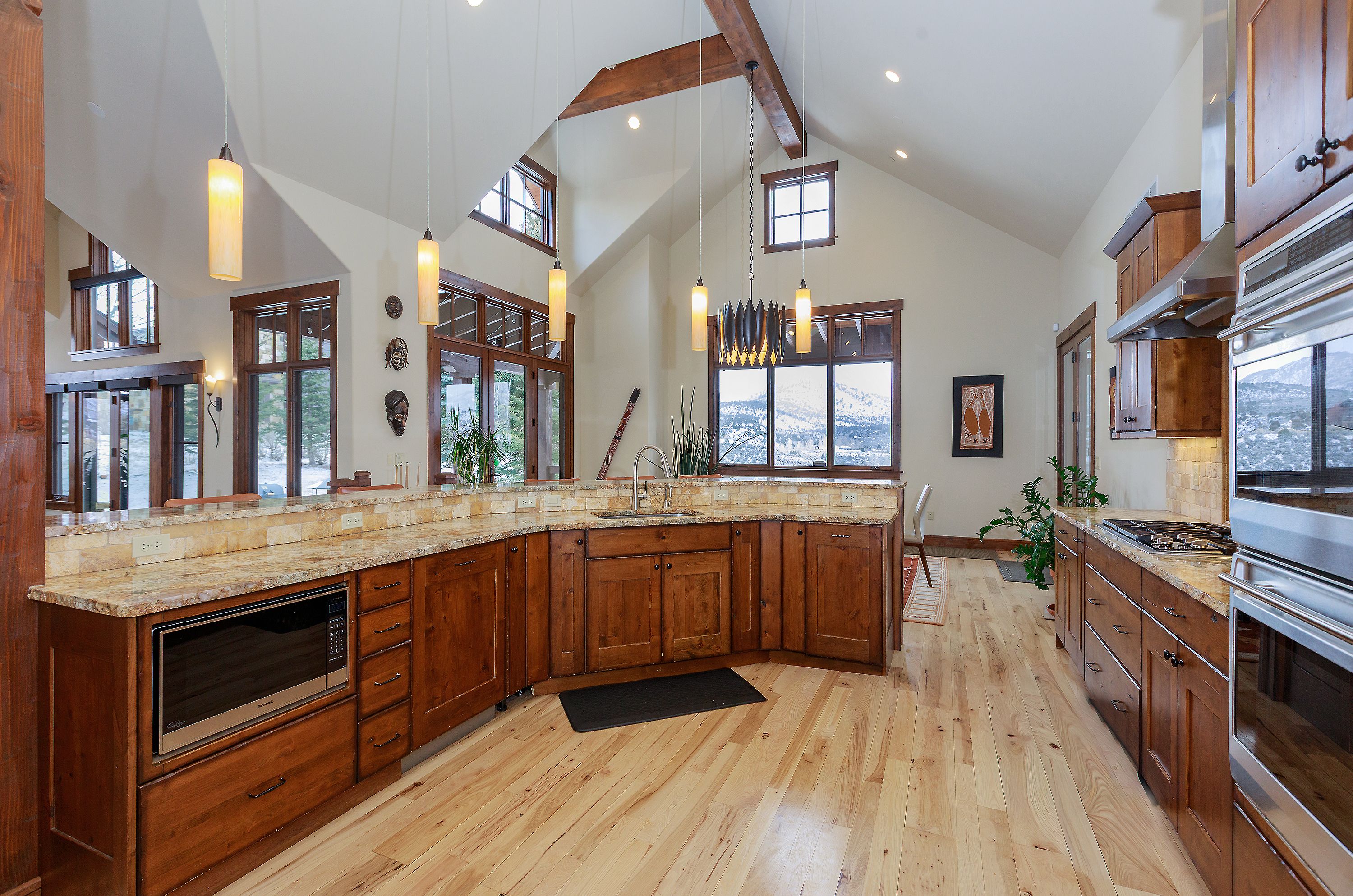 This spacious, rustic-style kitchen features warm wood cabinetry, granite countertops, and light-toned hardwood flooring under a dramatic vaulted ceiling with exposed beams. The layout includes a large corner sink area, a built-in microwave, and a professional-grade range, all illuminated by elegant pendant lighting. Large windows provide a scenic view of a snowy landscape, creating a cozy yet expansive atmosphere perfect for mountain living.