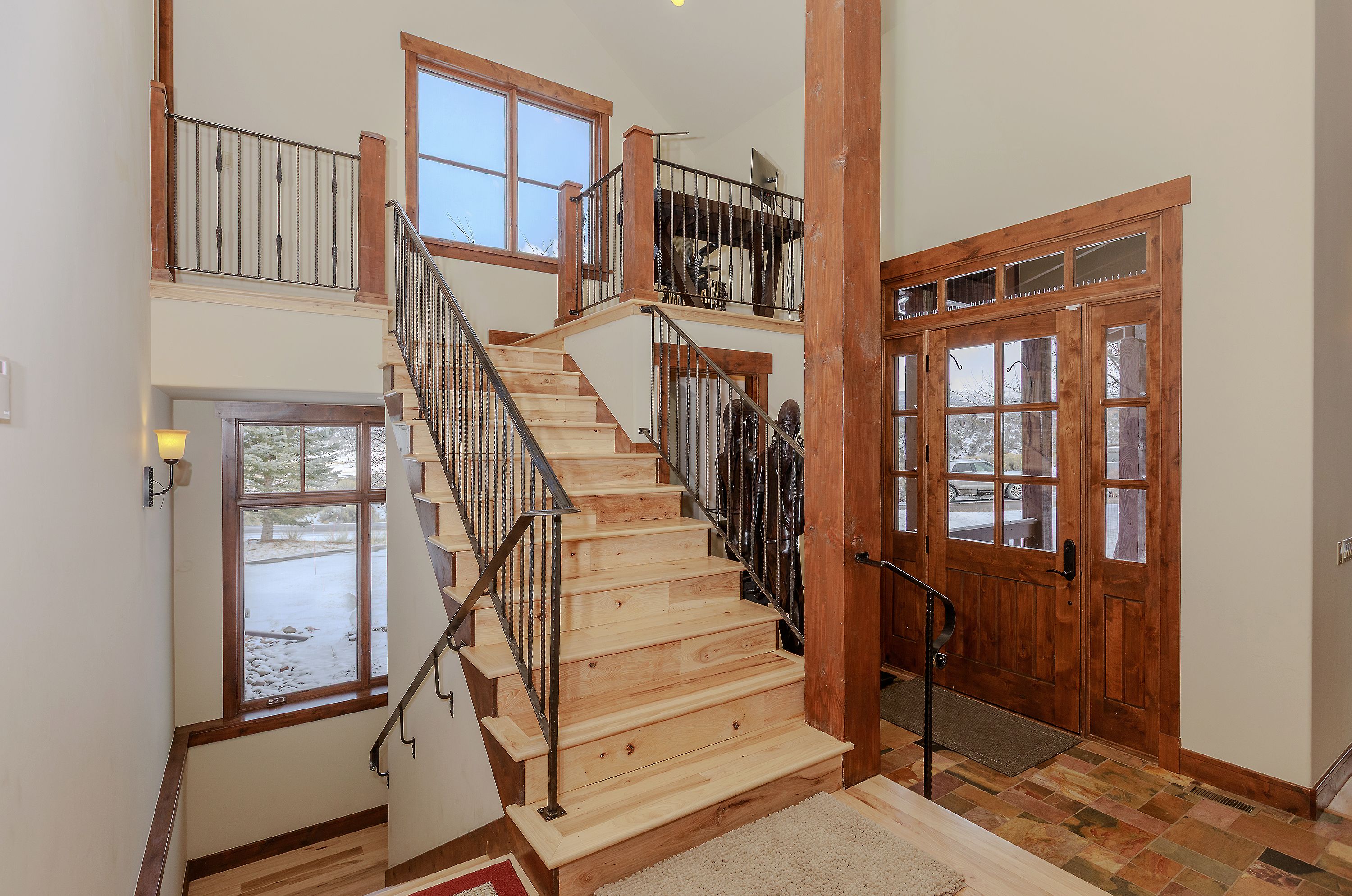 This grand entryway features a striking wooden staircase with wrought iron railings, serving as the focal point of the home's interior. The space is characterized by high ceilings, warm wood trim, and a large front door with glass panels that invite natural light into the foyer. The combination of rustic timber elements and stone-tiled flooring creates a welcoming, mountain-lodge aesthetic.