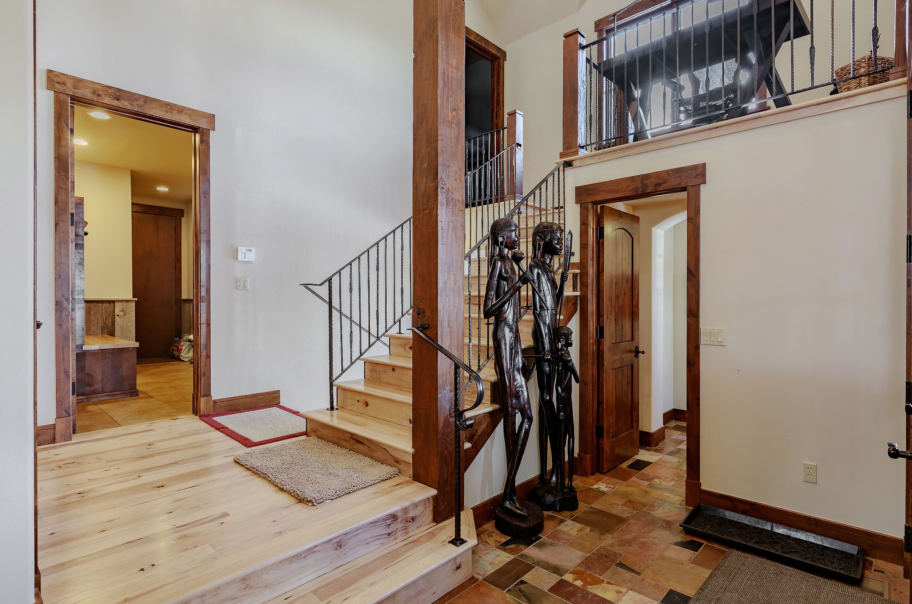This inviting entryway features a grand staircase with natural wood treads and ornate iron railings, anchored by a prominent timber support beam. The space is accented by striking, tall wooden sculptures and a mix of light hardwood and rustic slate tile flooring, creating a warm, mountain-lodge aesthetic. The perspective captures the transition between the foyer and the upper level, highlighting the home's architectural character and open, airy design.