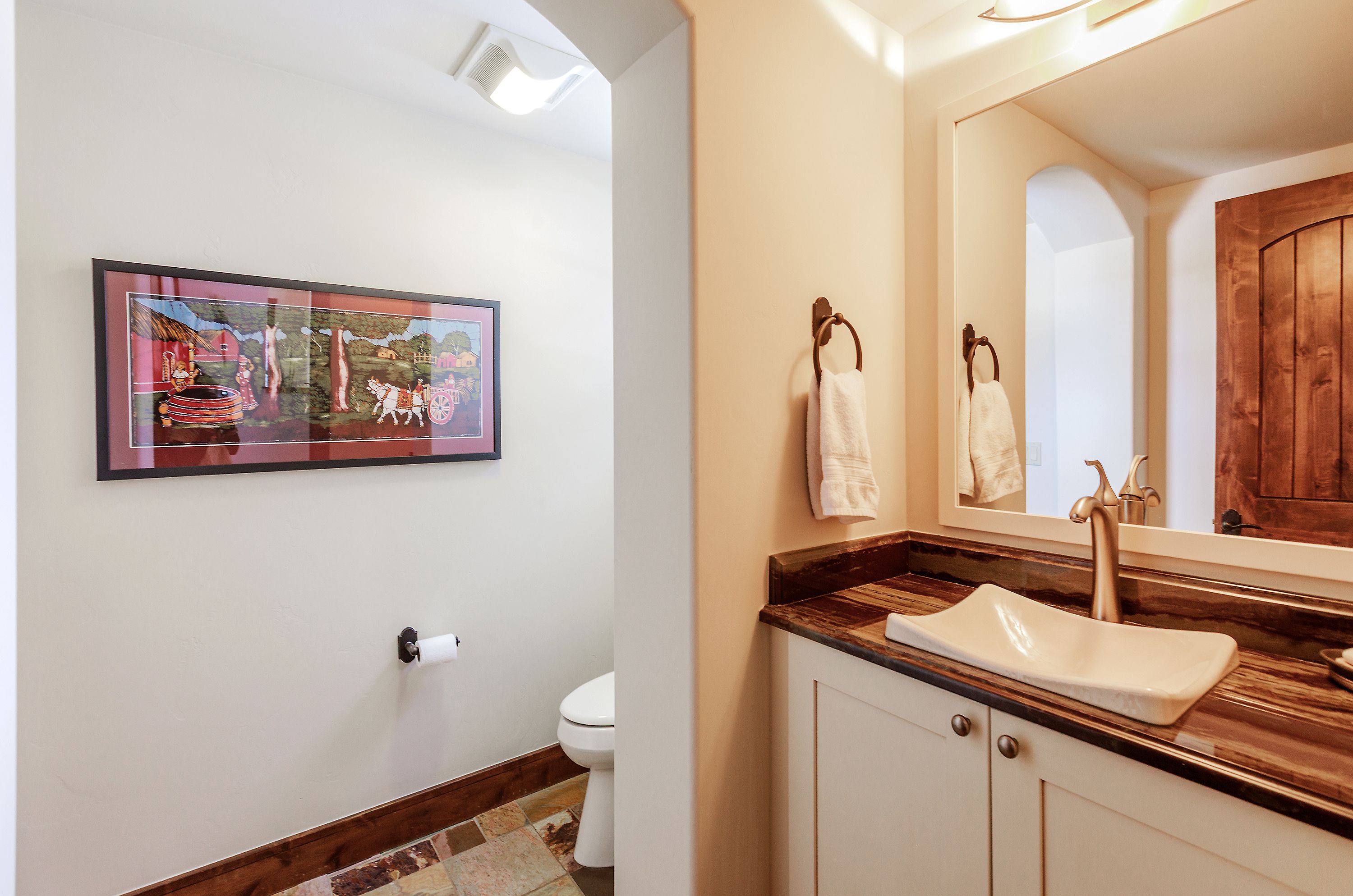 This bathroom features a unique, shallow vessel sink set atop a rich, dark wood-grain countertop, complemented by cream-colored cabinetry. A framed folk-art painting hangs on the wall leading into the water closet, while a large mirror reflects the warm, inviting lighting. The space combines rustic charm with functional design, creating a cozy and well-appointed guest bathroom.