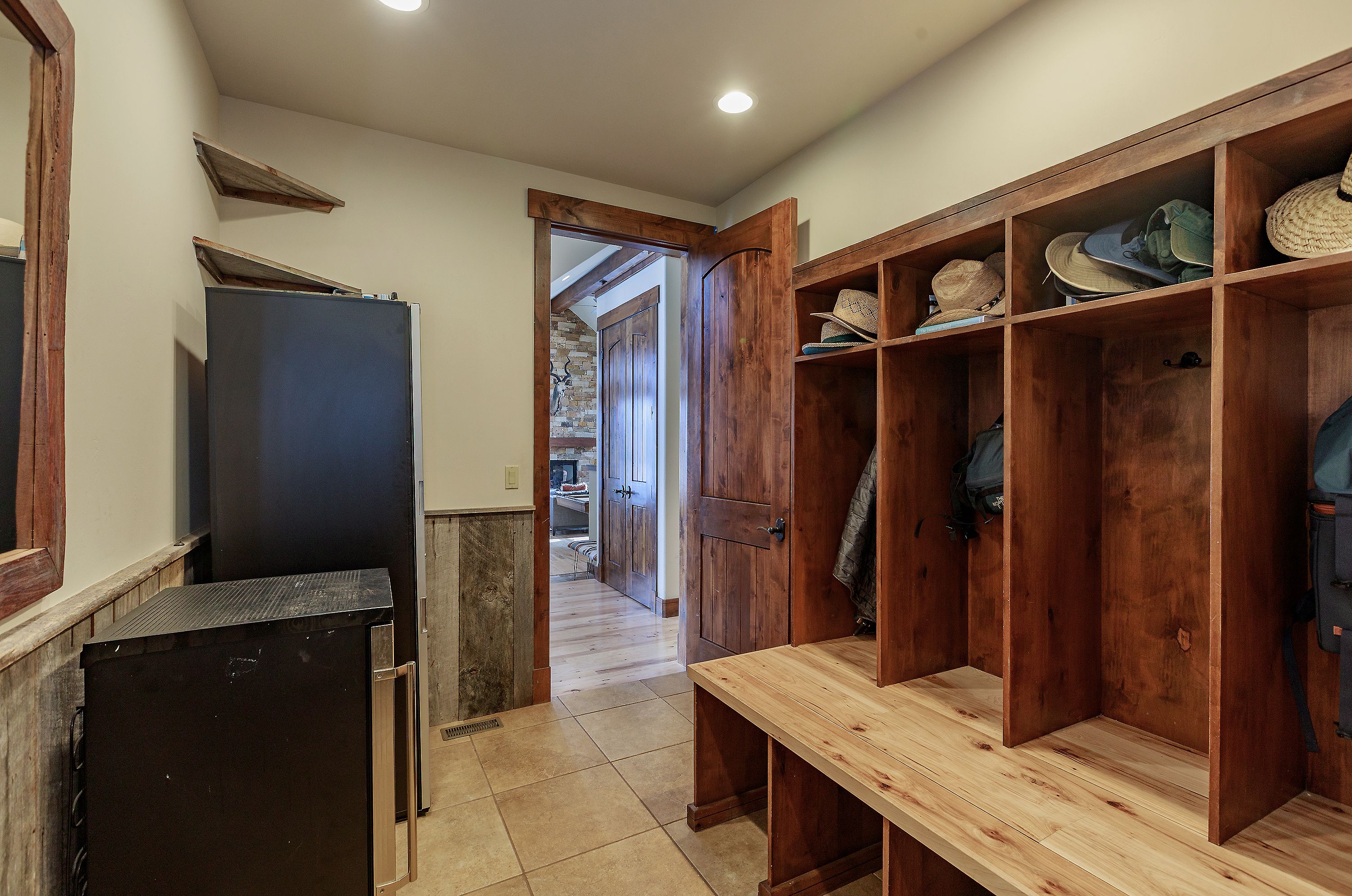 This mudroom area features custom built-in wooden lockers with integrated bench seating and overhead storage cubbies, perfect for organization. The space includes a small refrigerator, rustic wood paneling, and tile flooring, leading through a doorway into a cozy living area with a stone fireplace. The warm wood tones and functional design create a welcoming and practical transition space within the home.