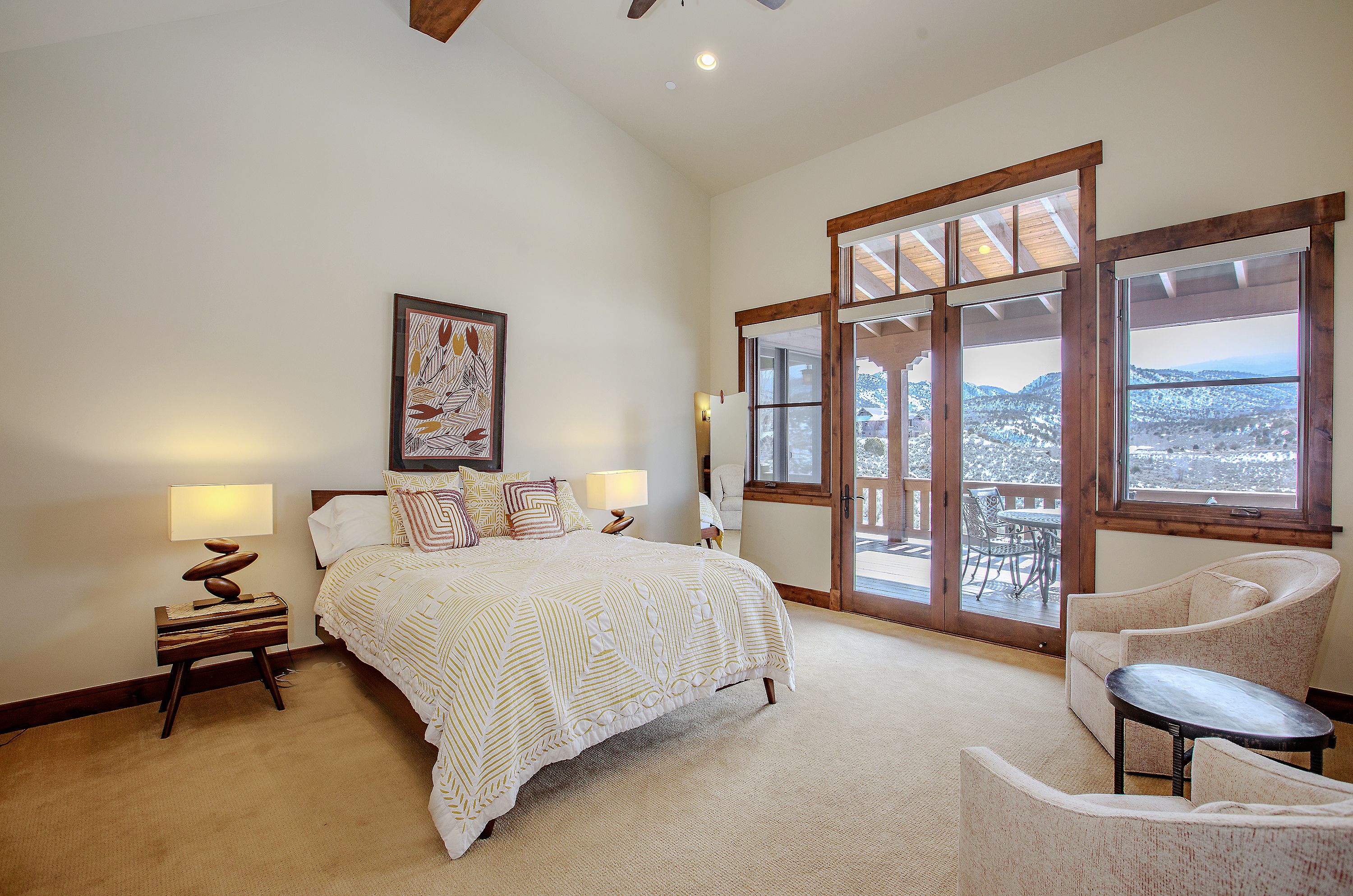 This spacious primary bedroom features a vaulted ceiling, neutral carpeting, and a large bed adorned with patterned bedding. A set of glass-paned French doors opens onto a private deck, offering stunning views of the snow-capped mountains beyond. The room is completed by two comfortable armchairs and a small side table, creating a serene and inviting retreat.