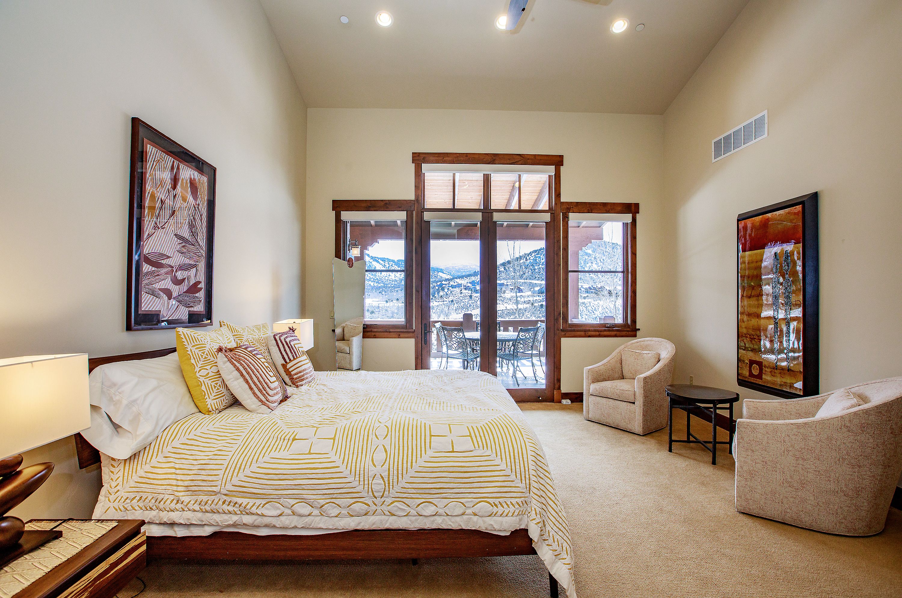 This spacious primary bedroom features a warm, neutral color palette with a large bed adorned in a geometric yellow and white quilt. The room is anchored by two comfortable armchairs and a small side table, while large windows and a glass door provide a stunning, unobstructed view of a snow-capped mountain landscape. The overall aesthetic is serene and inviting, blending rustic wood accents with contemporary decor.
