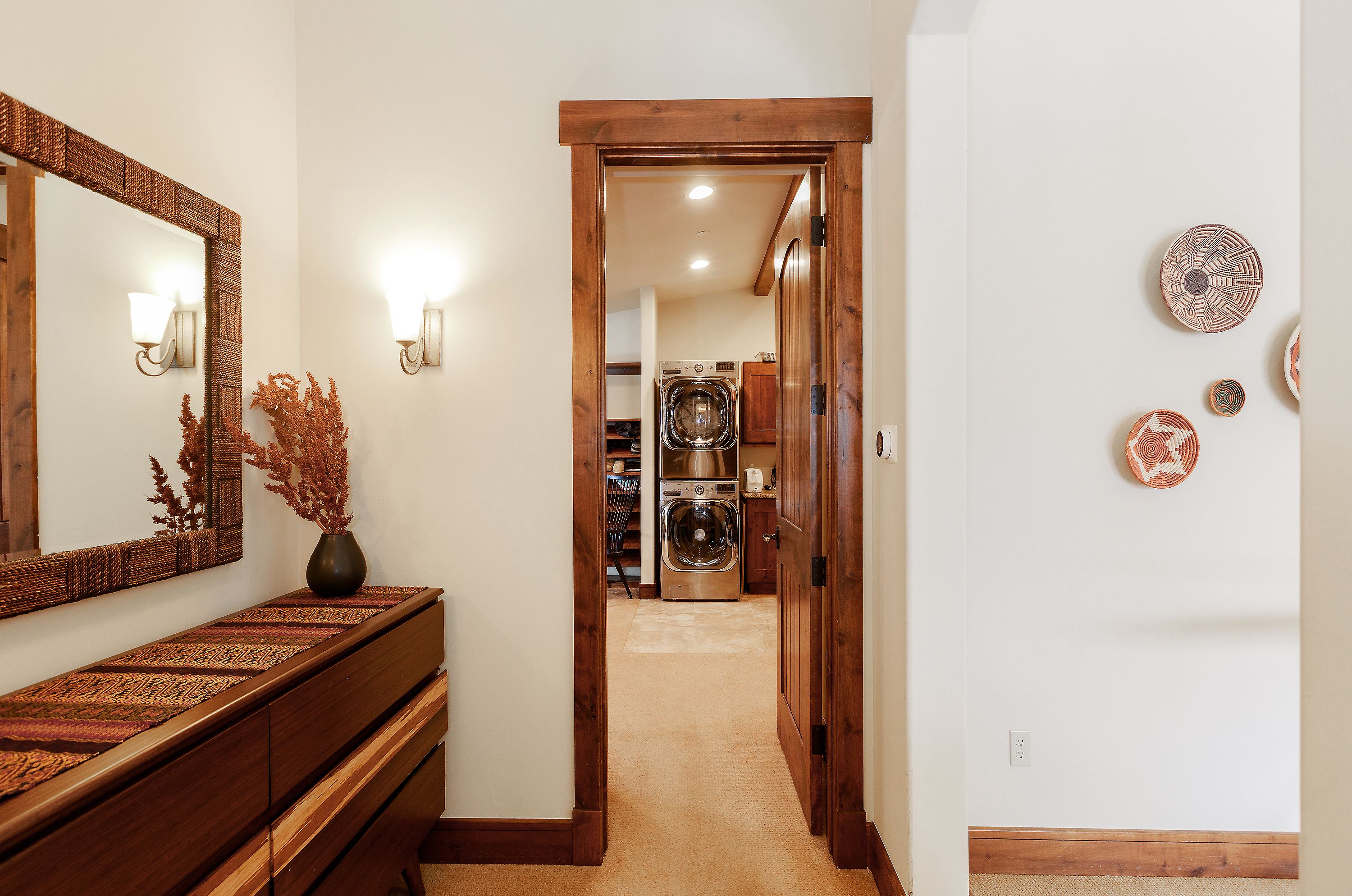 This interior view captures a warm, rustic-style hallway leading into a laundry room. The foreground features a wooden console table topped with a woven mirror and decorative dried botanicals, while the wall to the right is adorned with circular woven wall art. The perspective draws the eye through the wooden door frame toward the stainless steel laundry appliances, creating a sense of depth and cozy, well-maintained living space.