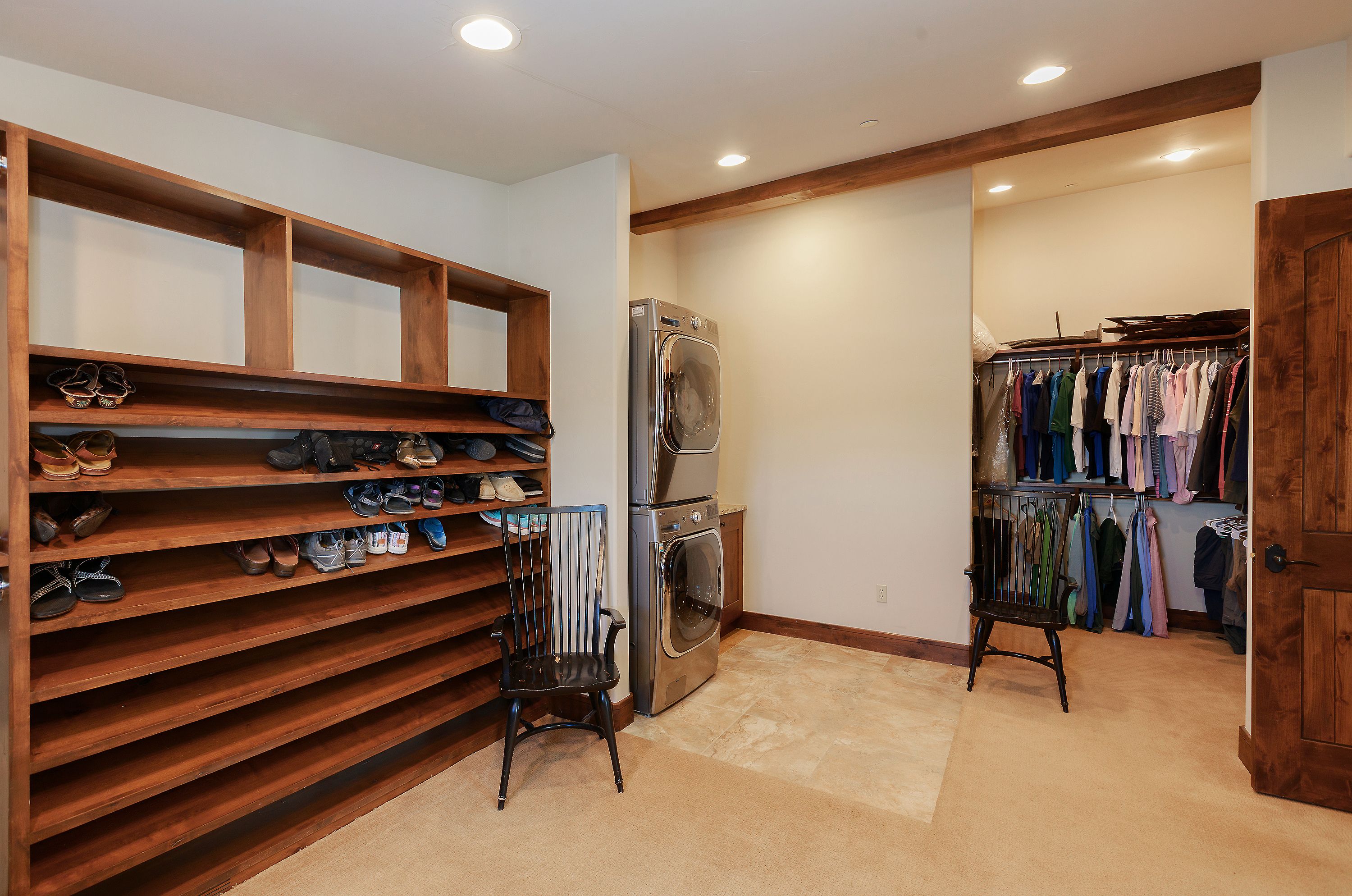 This functional laundry room features a stacked washer and dryer unit set against a neutral wall, complemented by extensive custom wooden shelving for shoe storage. The space includes a walk-in closet area with hanging racks and a pair of classic black spindle chairs, creating a practical and organized mudroom-style environment. The room is finished with a combination of carpet and tile flooring, illuminated by recessed ceiling lights.