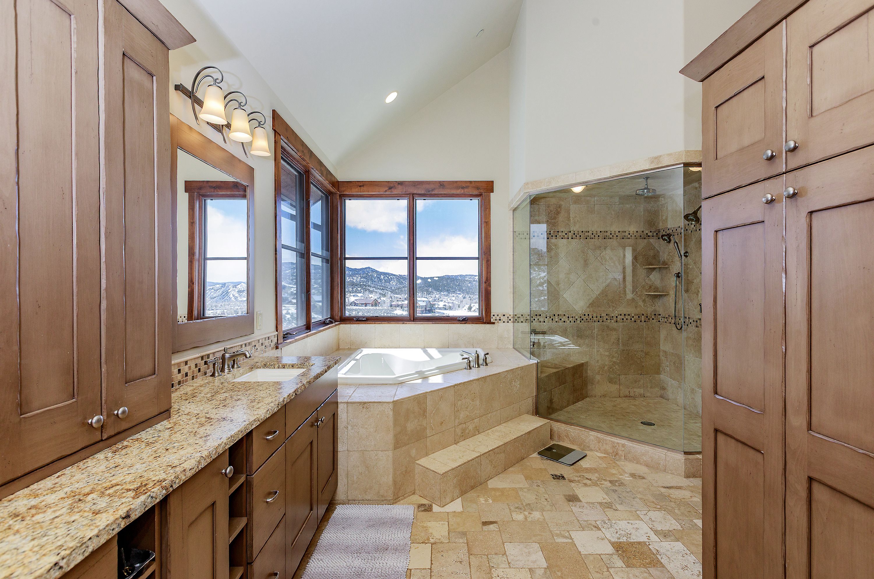 This spacious primary bathroom features a luxurious corner soaking tub set against a large window with scenic mountain views. The room is appointed with warm-toned wooden cabinetry, granite countertops, and a walk-in glass-enclosed shower with tiled walls. The overall aesthetic is rustic and elegant, utilizing natural stone flooring and high ceilings to create an airy, inviting atmosphere.