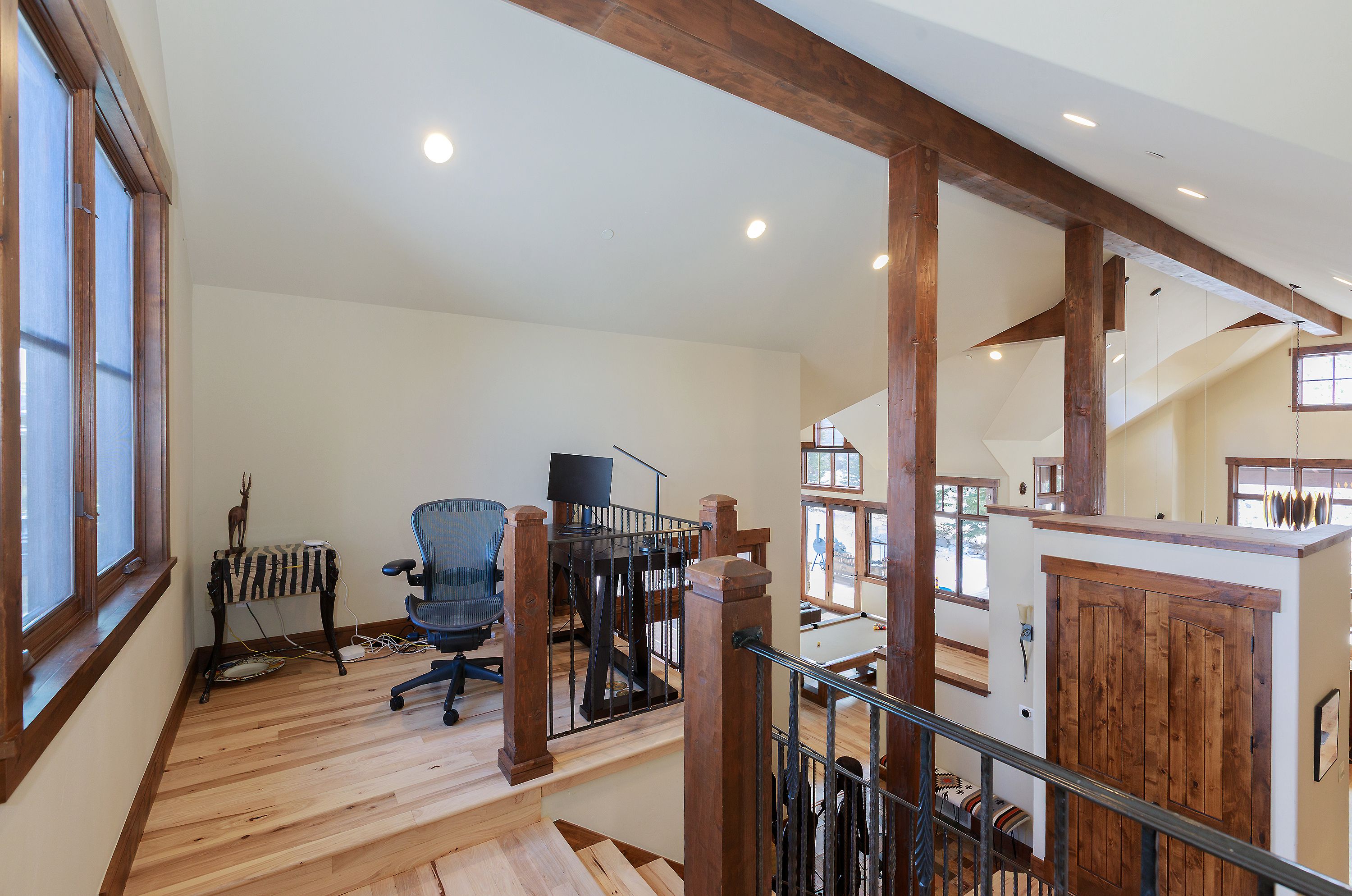This elevated home office loft overlooks a lower living area, featuring a rustic-modern aesthetic with exposed timber beams and light hardwood flooring. The space is furnished with a sleek ergonomic office chair and a dark wood desk, framed by a decorative metal railing that adds architectural character. The open-concept design creates a bright, airy atmosphere that seamlessly connects the workspace to the rest of the home.