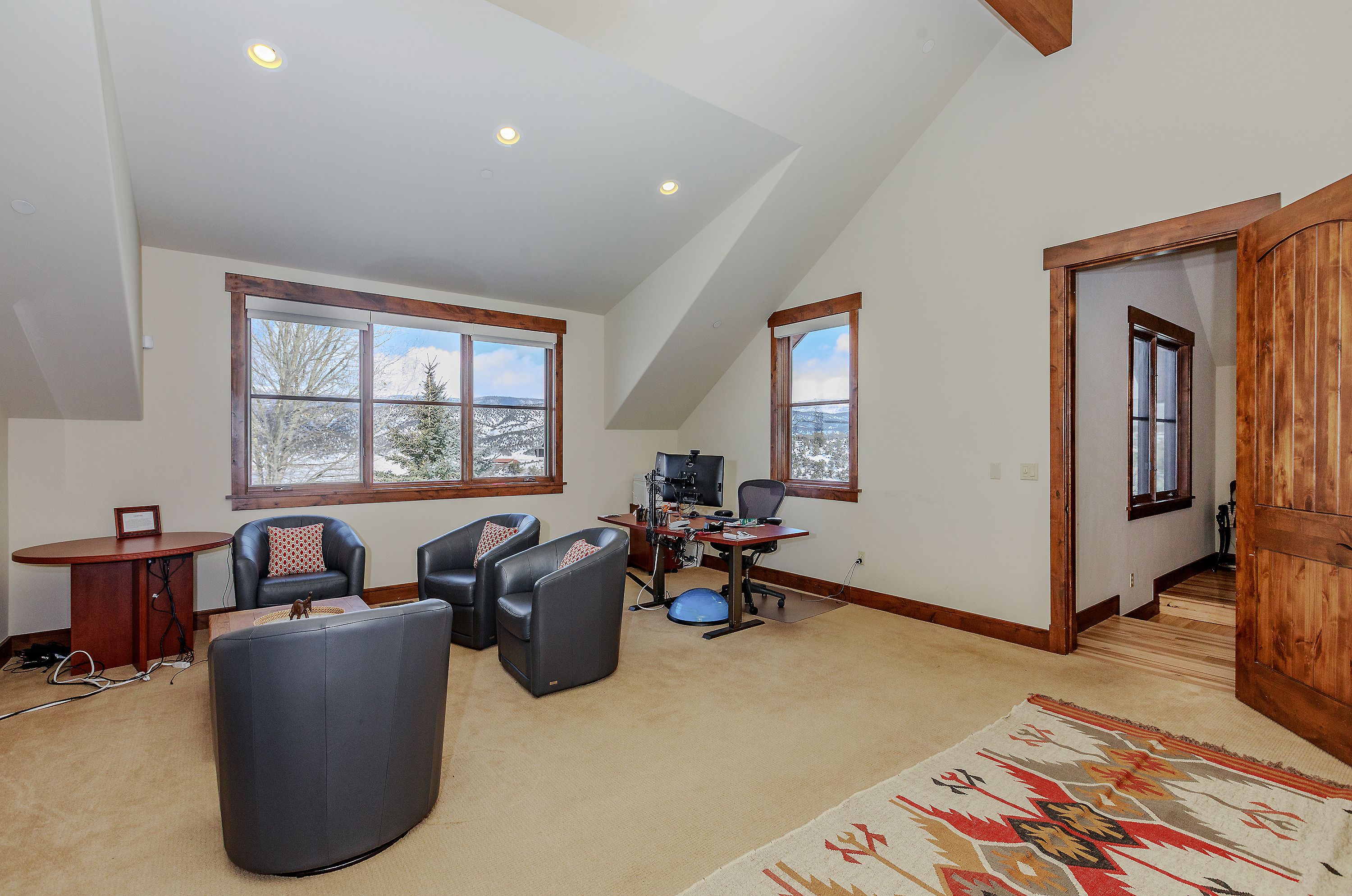 This spacious home office features a vaulted ceiling with exposed wood beams and large windows that offer scenic mountain views. The room is furnished with a professional desk setup, a comfortable seating area with leather armchairs, and a patterned area rug, creating a versatile workspace. The warm wood trim and neutral color palette provide a cozy yet functional atmosphere suitable for a productive home environment.