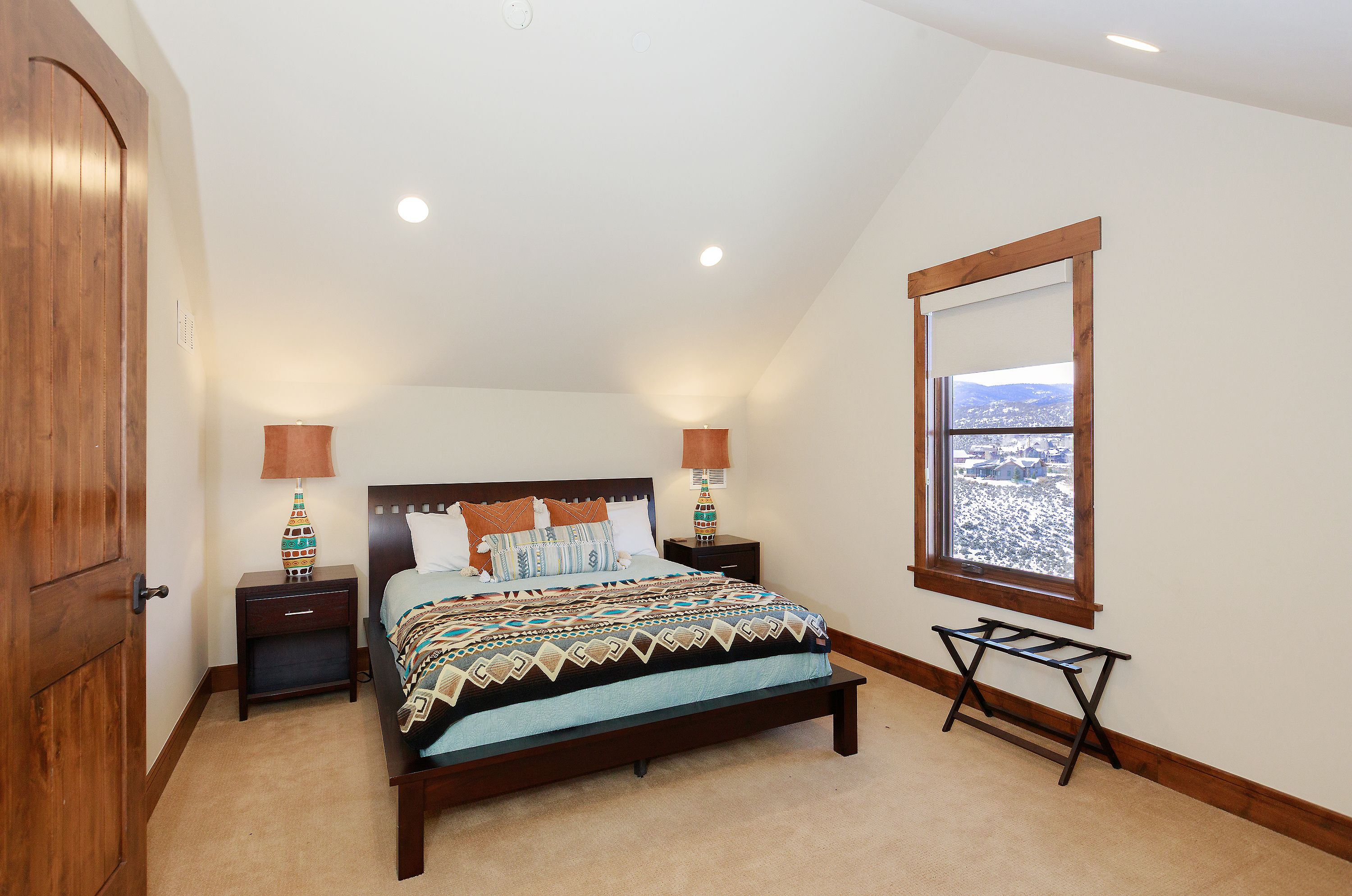 This cozy guest bedroom features a dark wood bed frame adorned with a patterned, southwestern-style quilt and decorative pillows. The room is illuminated by two bedside lamps with colorful, patterned bases and recessed ceiling lights, while a large window provides a scenic view of a snowy landscape. The space is finished with warm-toned carpeting and wood trim, creating a comfortable and inviting atmosphere.