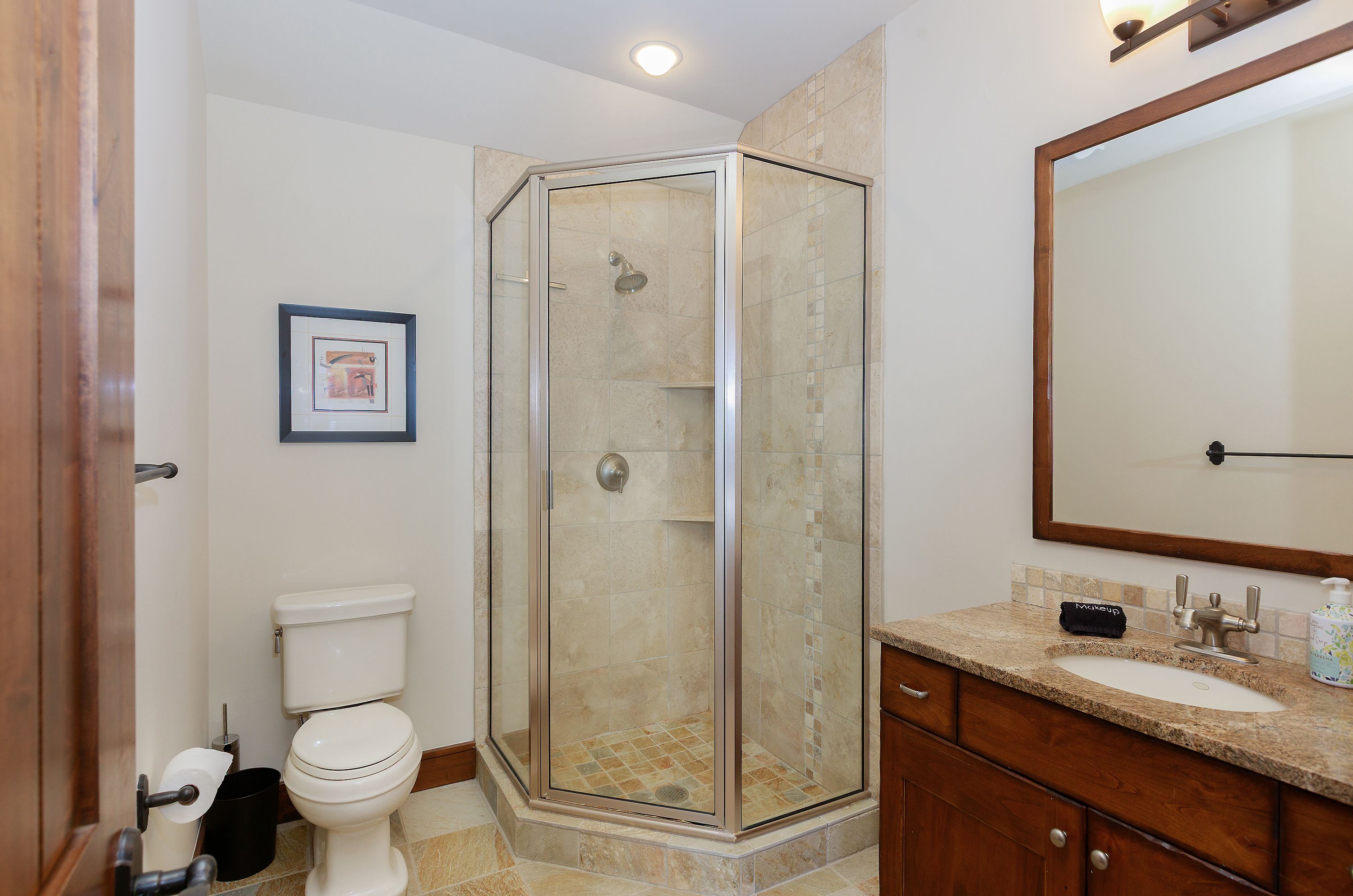 This compact, functional bathroom features a corner glass-enclosed shower with neutral-toned tile work and a matching stone-topped vanity with a dark wood cabinet. The space is well-lit with recessed lighting and a wall-mounted mirror, creating a clean and practical aesthetic. The perspective is a straight-on view that captures the efficient layout of the room, including the toilet and vanity area.