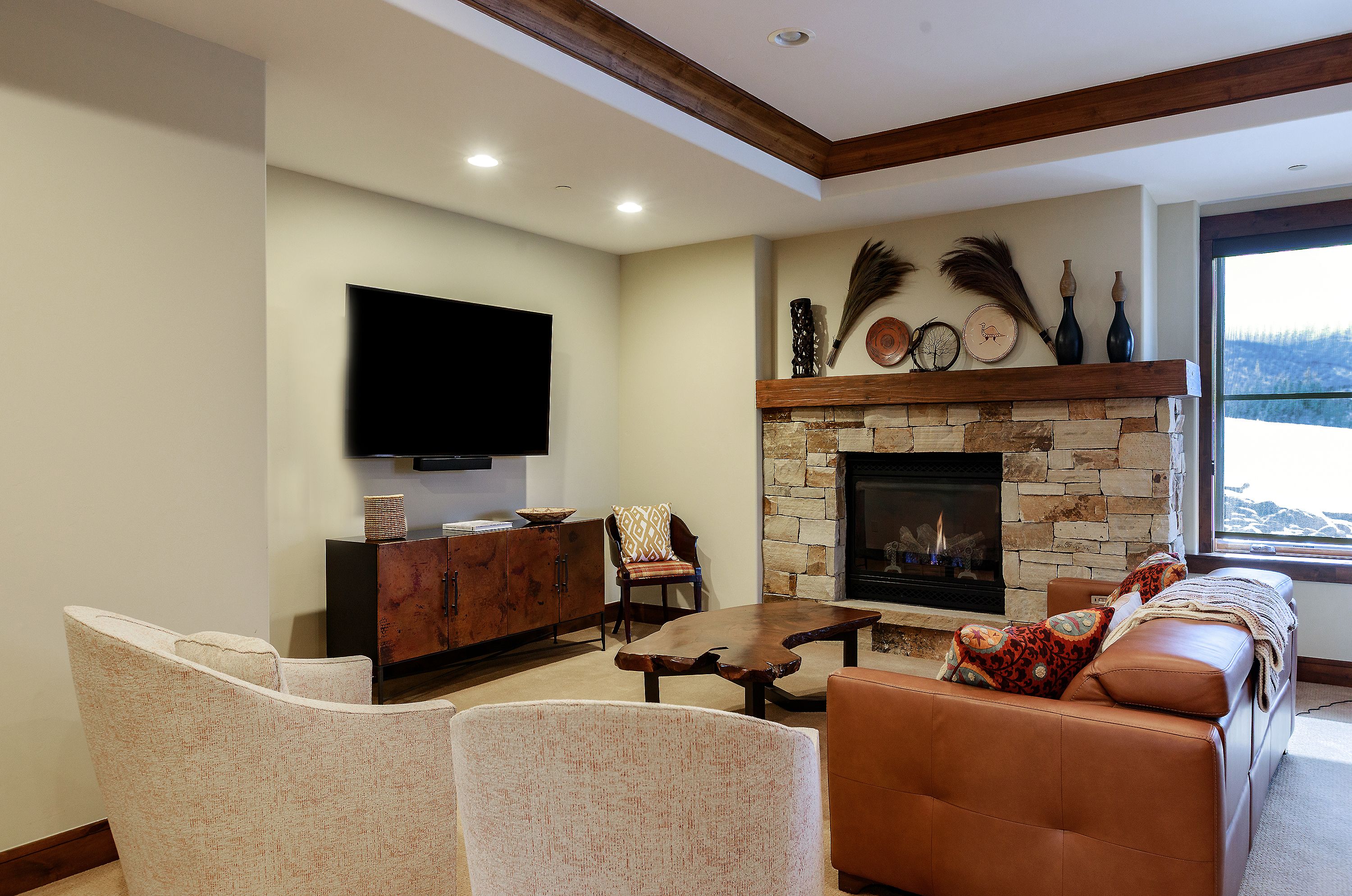 This cozy living room features a rustic stone fireplace as its focal point, complemented by a warm, earthy color palette and natural wood accents. The space is furnished with a comfortable leather sofa, two light-toned armchairs, and a unique live-edge coffee table, creating an inviting atmosphere. A large flat-screen TV is mounted above a dark wood media console, while a window to the right provides a view of a snowy landscape, emphasizing the home's mountain-inspired aesthetic.