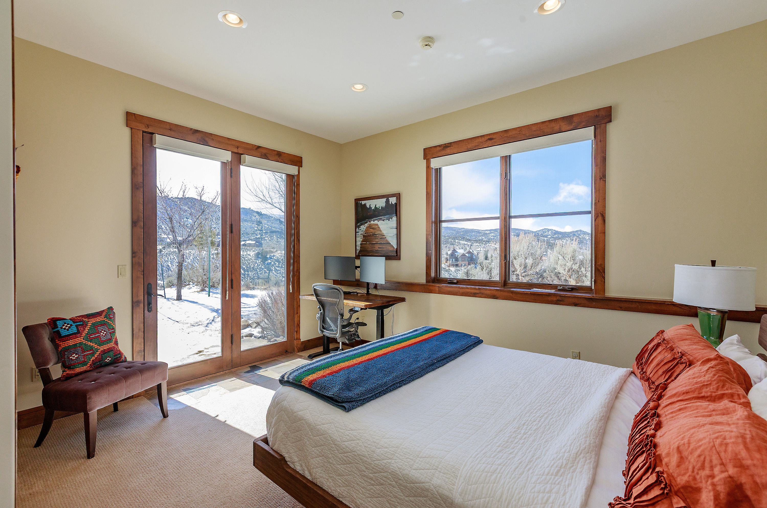 This cozy bedroom features a comfortable bed with a vibrant, multi-colored striped throw blanket and textured orange pillows, creating a warm and inviting atmosphere. A dedicated workspace with dual monitors and an ergonomic chair is positioned near a large window and glass doors that offer expansive, scenic views of a snow-dusted landscape. The room is accented by a plush, dark-toned armchair with a patterned throw pillow, blending rustic charm with modern functionality.