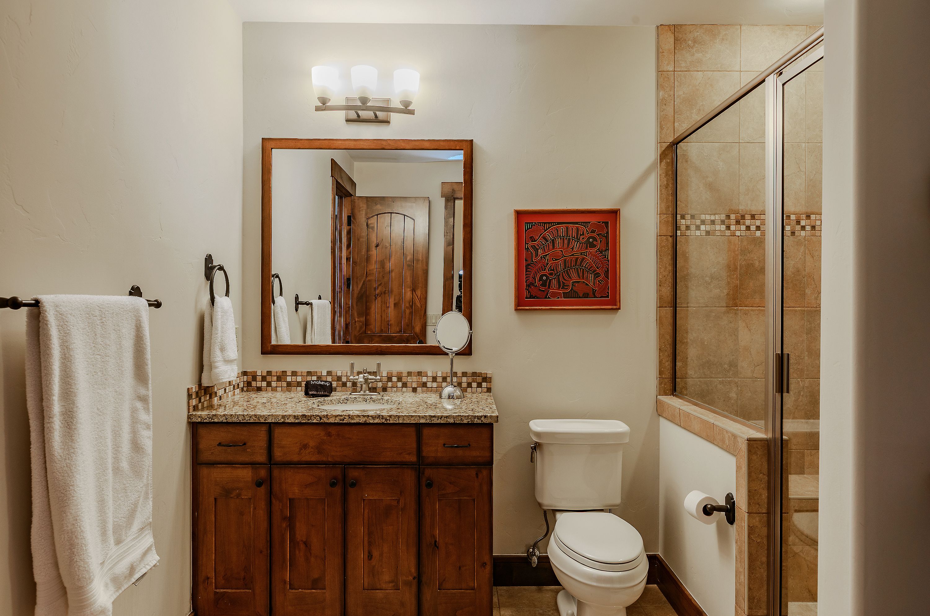 This well-appointed bathroom features a rustic wooden vanity topped with granite, complemented by a framed mirror and a three-light fixture. The space includes a tiled walk-in shower with a glass door and a standard toilet, all tied together by a warm, earthy color palette. The perspective is a straight-on shot that highlights the functional layout and cozy, traditional aesthetic of the room.
